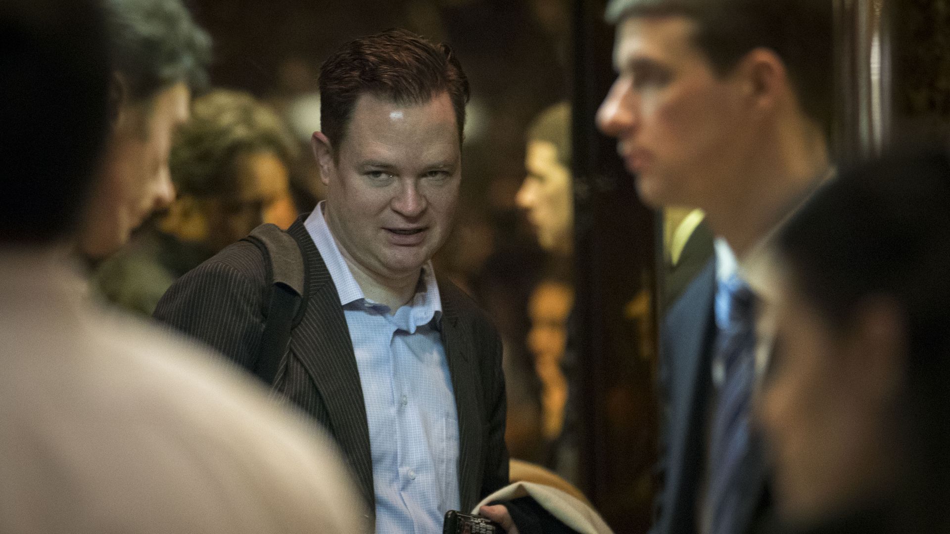 Man in a light blue shirt and dark striped blazer holding a phone, talking to three blurred people in a dimly lit indoor setting with warm tones.