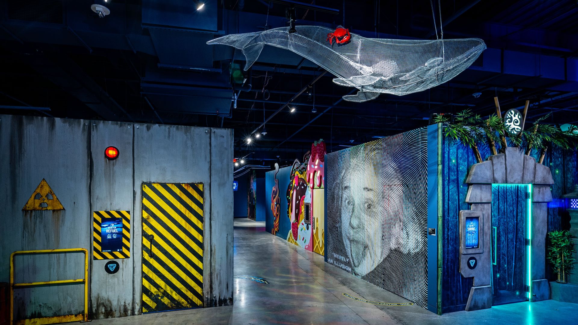 Indoor corridor with a gray wall featuring a yellow-black striped door, a radiation sign, and a red light. Opposite a blue-lit entrance with plants and a digital screen, and a netted whale sculpture overhead.