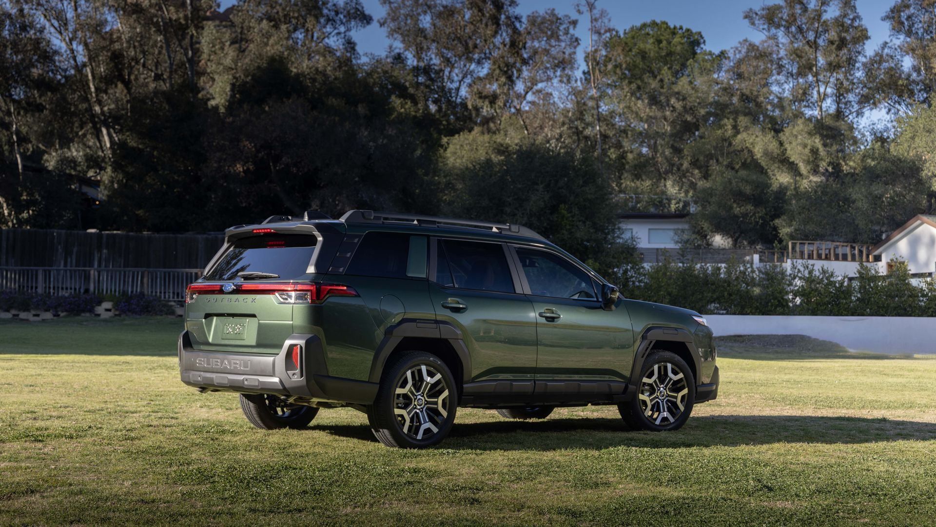 Rear and side view of a green Subaru Outback SUV parked on a grassy lawn, with tall trees and a blue sky in the background; roof rails visible, fence and houses behind.