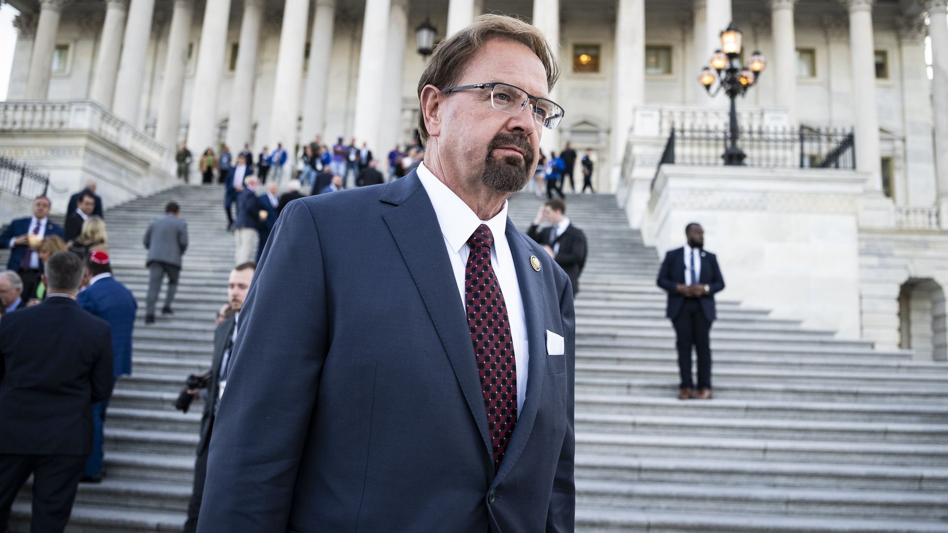 A man in a dark blue suit with a red patterned tie and glasses stands on grand stone steps in front of a neoclassical government building with tall white columns; a crowd is on the steps behind him.
