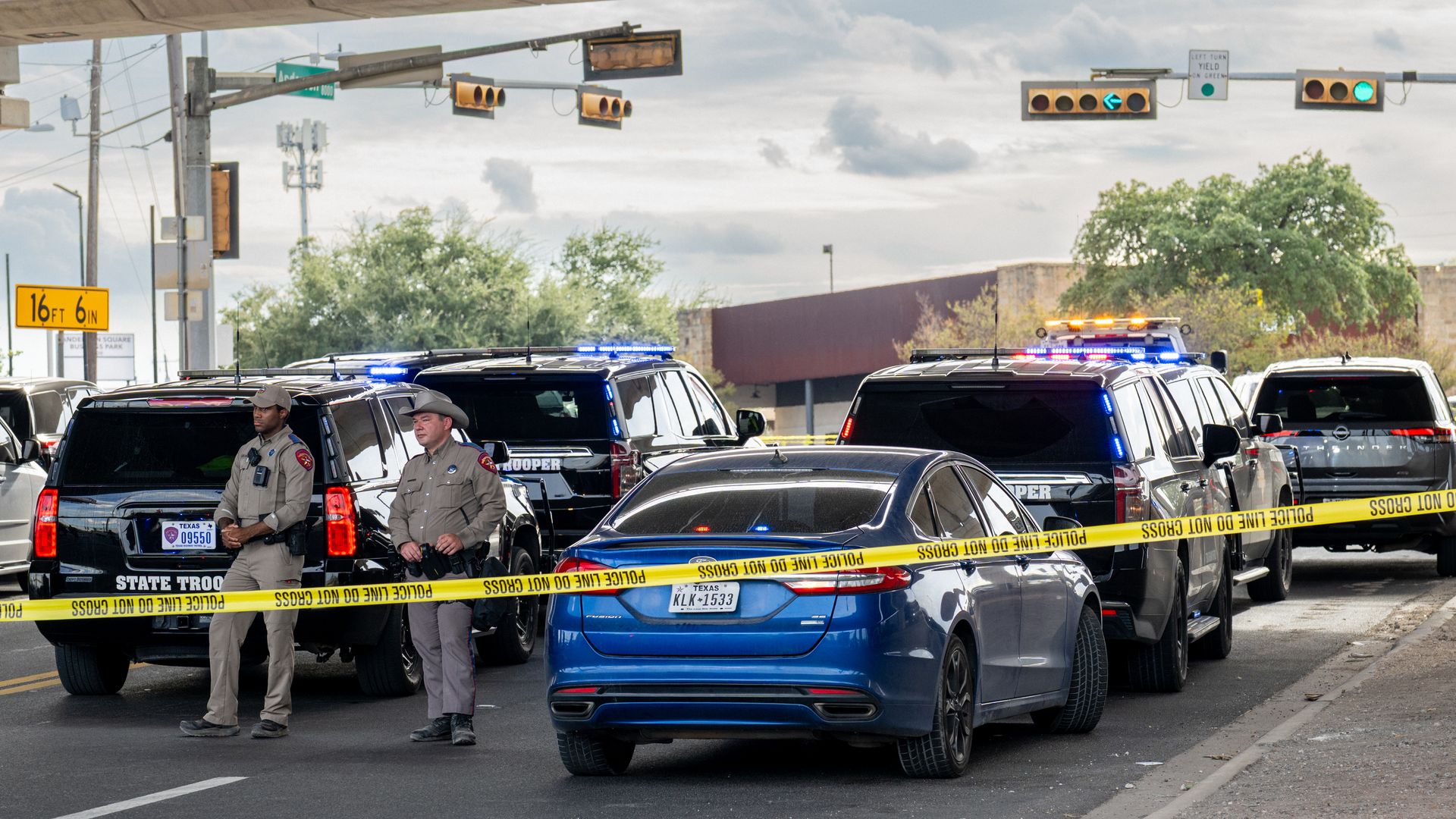 Under a concrete overpass, police SUVs with flashing lights block the street. Yellow "POLICE LINE DO NOT CROSS" tape cordons the scene as two officers stand in the foreground.