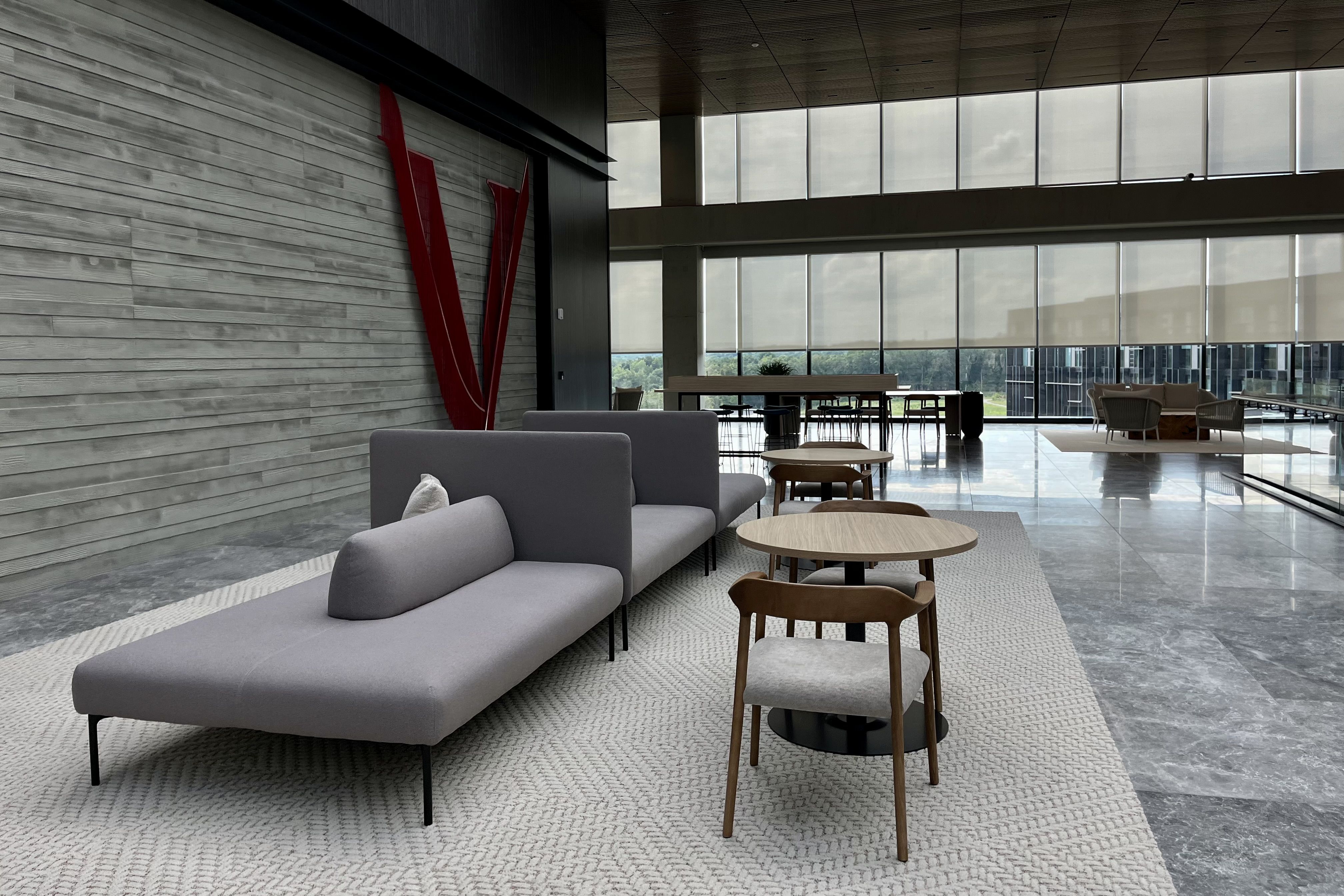 Modern spacious waiting area with gray sectional sofas, light wooden tables and chairs on a textured carpet, large floor-to-ceiling windows with blinds, and a red sculpture on gray wood wall.