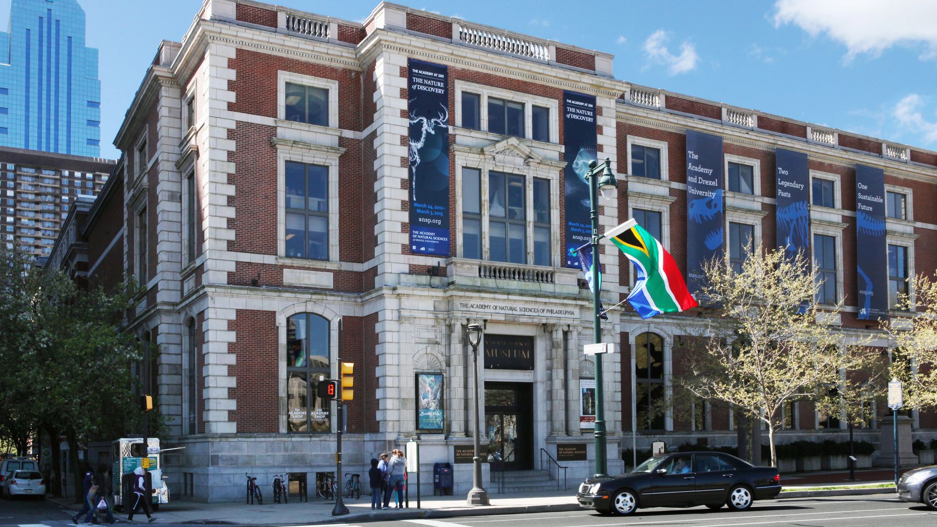 Brick and stone building of The Academy of Natural Sciences of Philadelphia with blue banners about an exhibit, a South African flag on a lamp post, and a crosswalk in front.