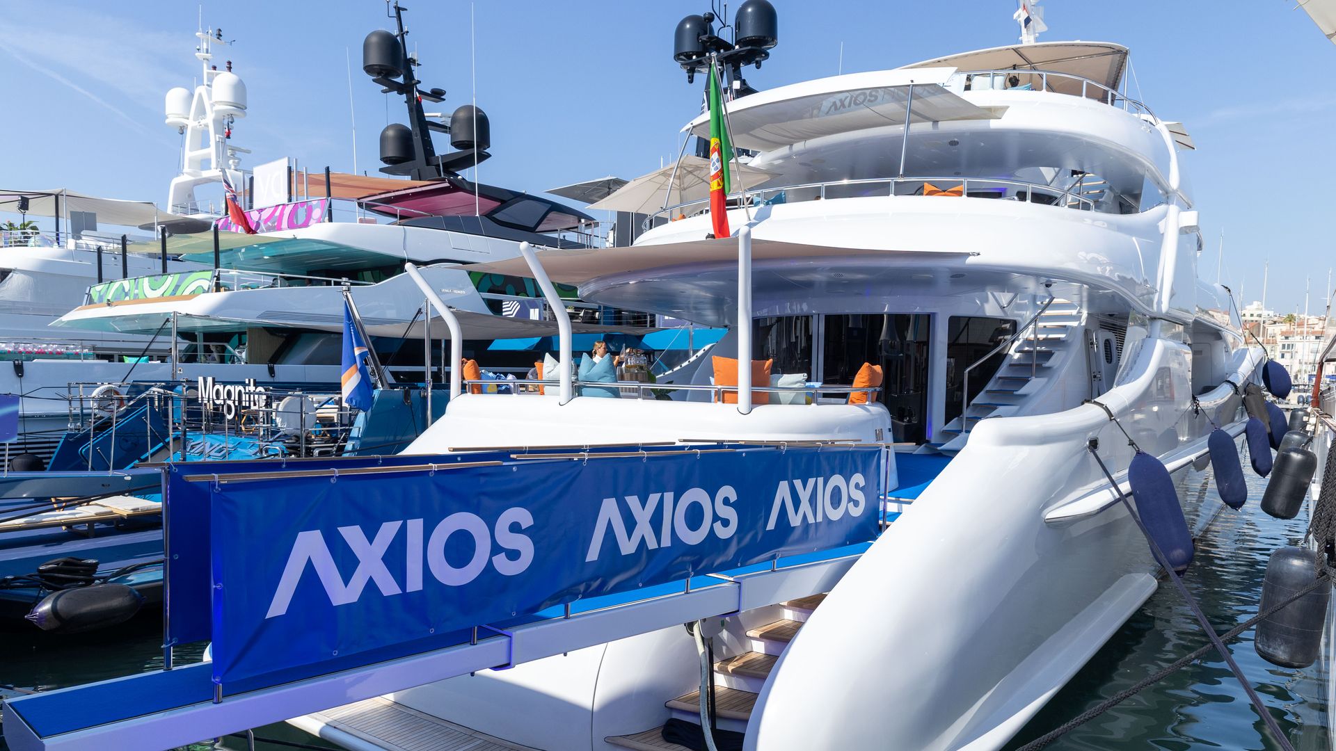 White luxury yachts docked at a marina under a clear blue sky; blue Axios banners along the pier, orange cushions on seating, and a Portuguese flag on one boat.