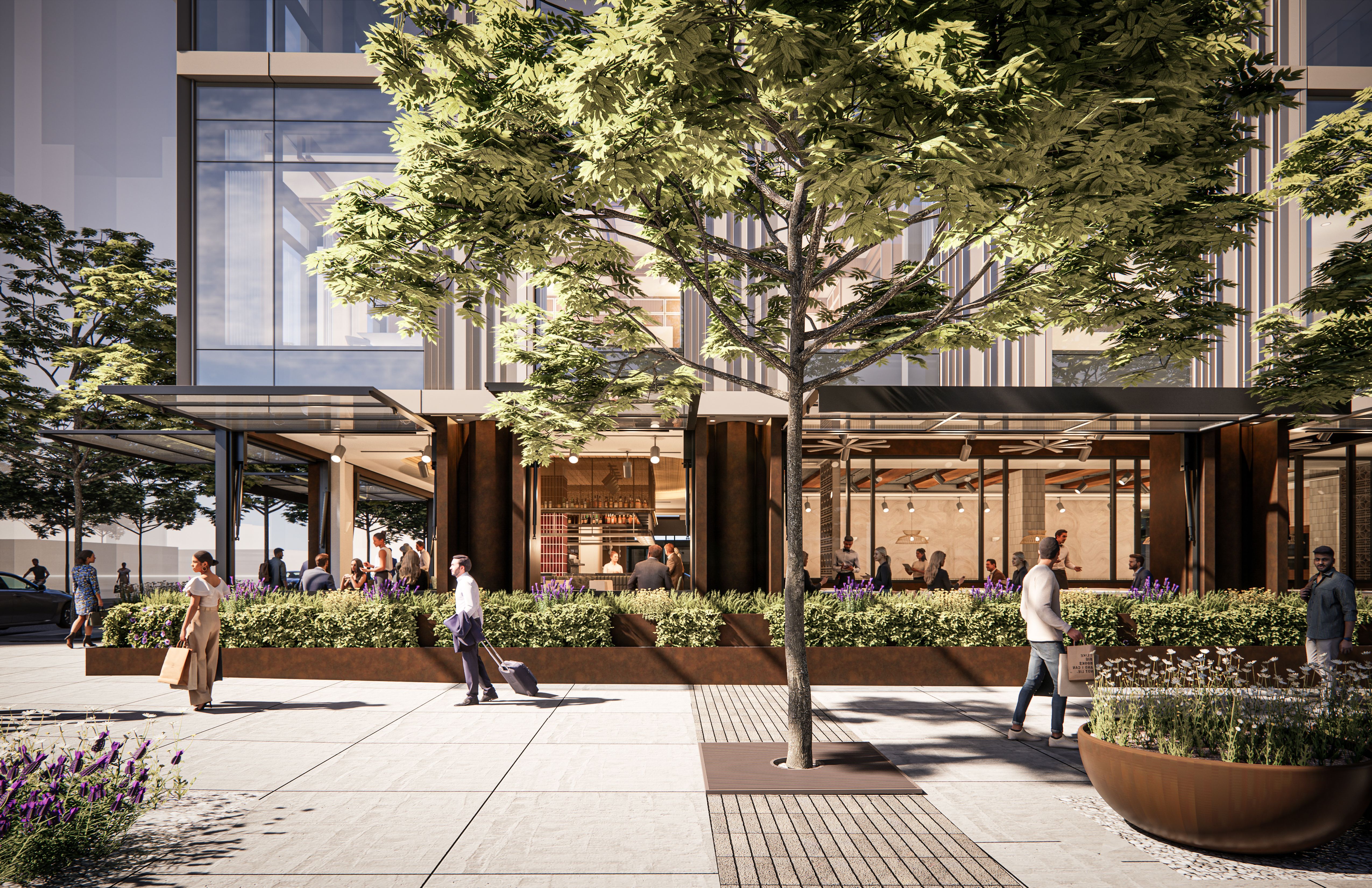 Urban plaza with a large tree in front of a modern glass-front building. Ground-floor shops, people walking, and planters with purple flowers and outdoor seating.