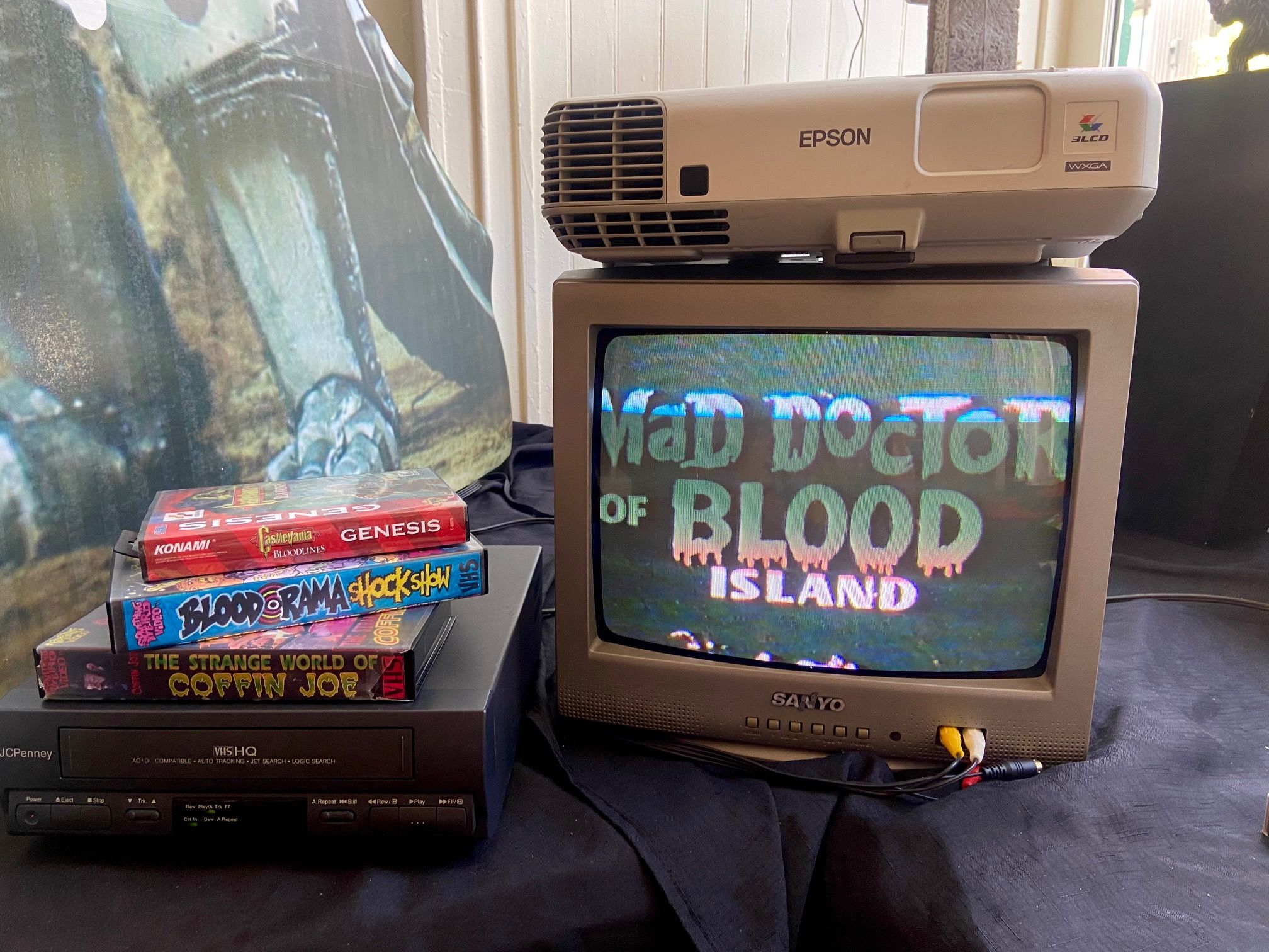Vintage media setup: beige Epson projector atop a CRT TV, with a stack of retro game boxes (Castlevania: Bloodlines) and a VCR below. The TV screen shows distorted text resembling ISLAND.