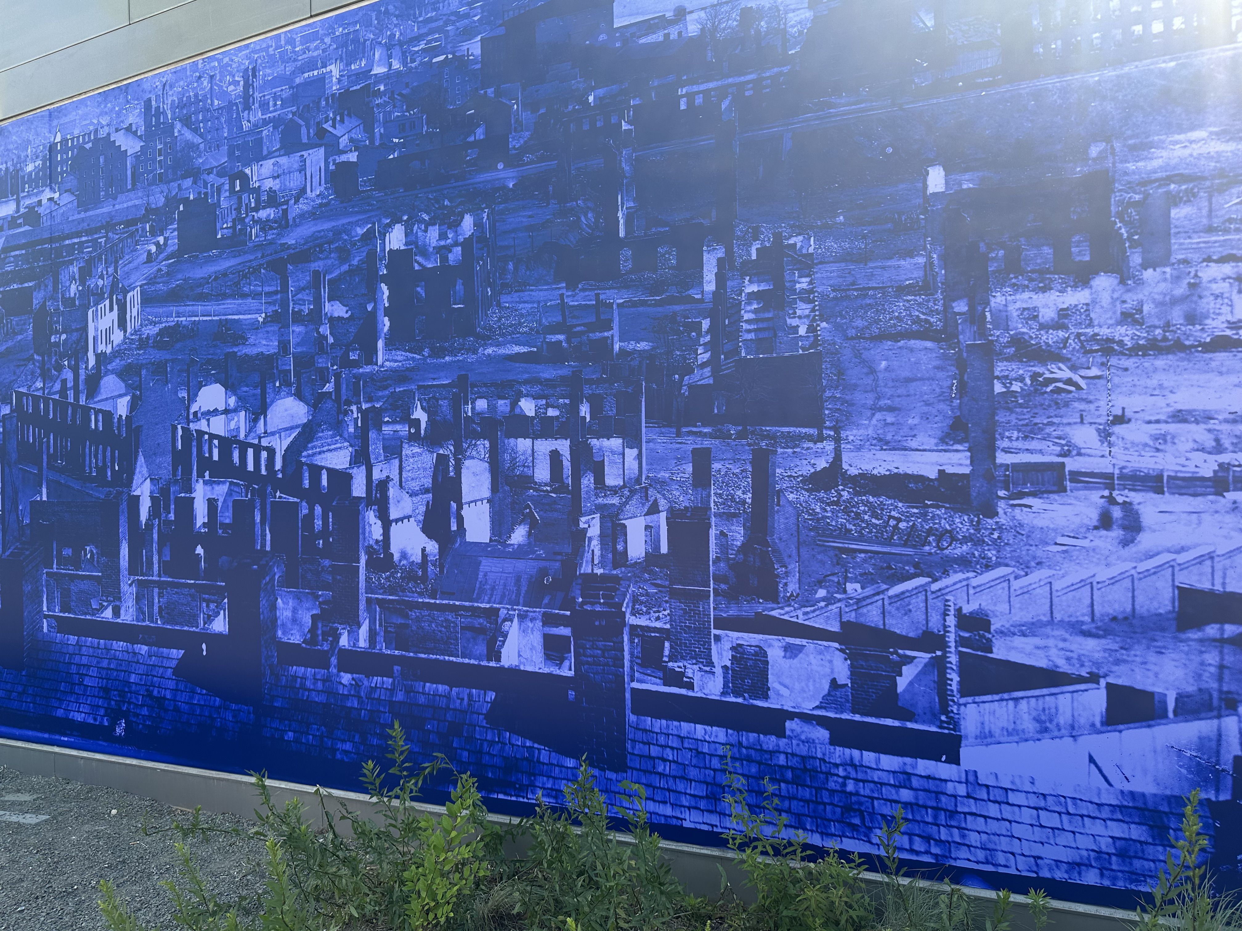 Blue-toned mural of a dense cityscape with numerous buildings and rooftops in layered silhouettes. Foreground shows gravel ground and green plants along the base.