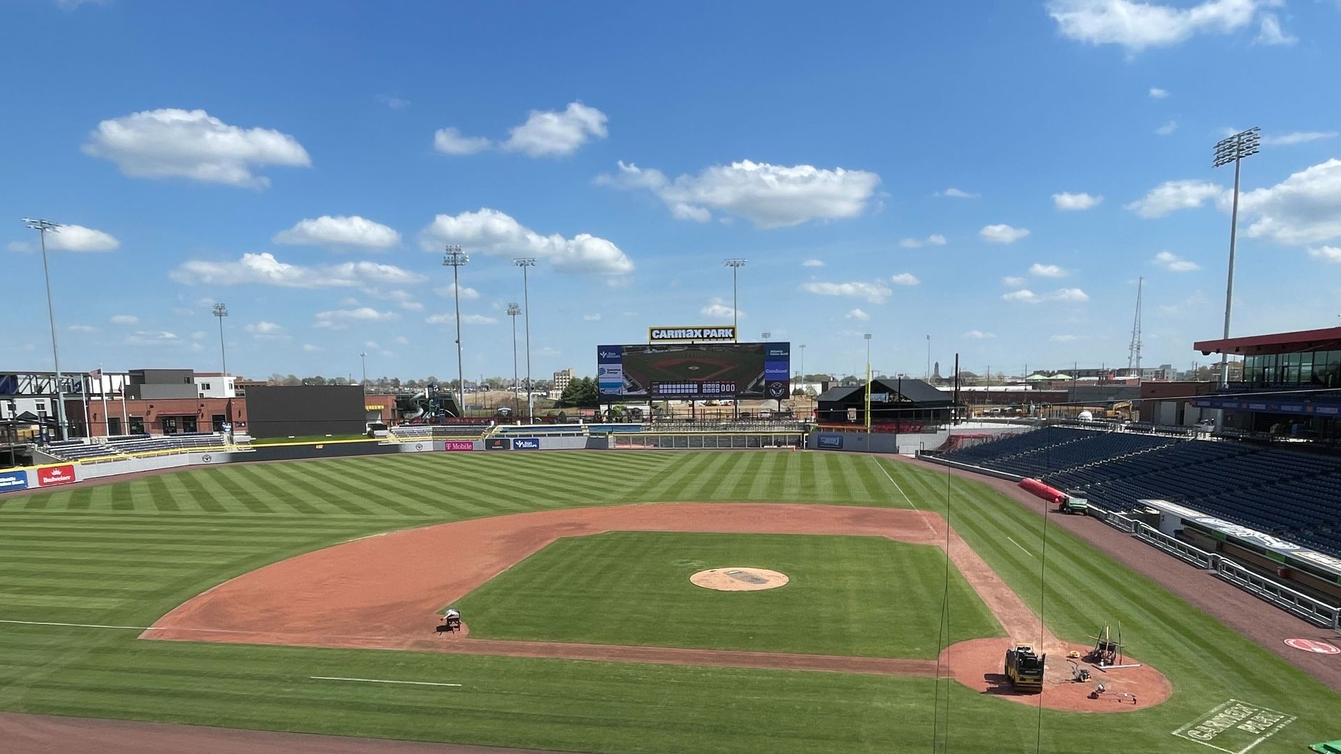 Wide view of CarMax Park baseball stadium on a sunny day: green field, brown infield, large center scoreboard, empty blue seats, tall light towers, and a few workers near the right field area.