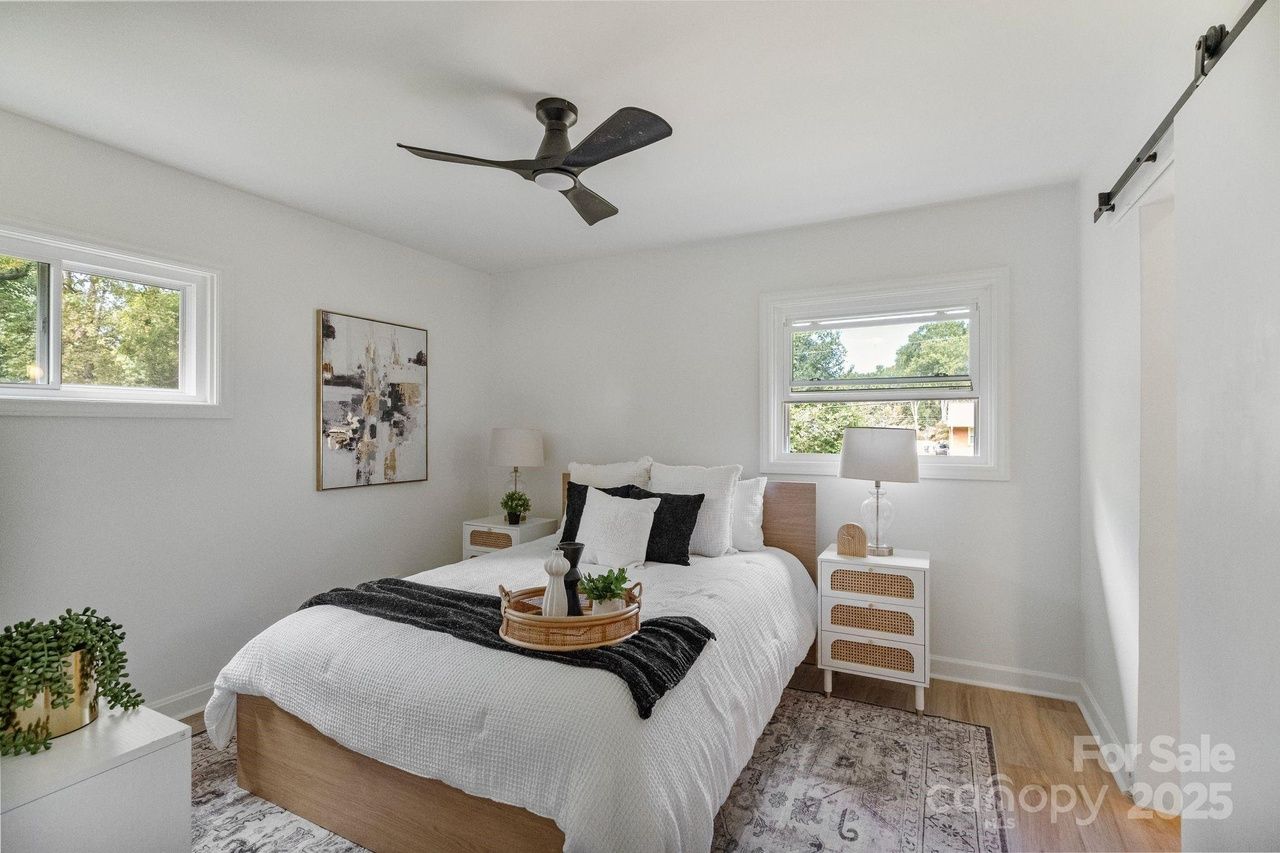 Bright bedroom with white walls, wooden bed with white bedding and black pillows, two white nightstands with lamps, ceiling fan, windows showing greenery outside, and abstract art on wall.