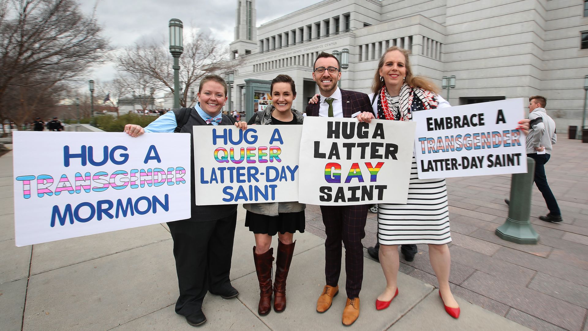 Four people on a plaza hold signs that read: "Hug a transgender Mormon," "Hug a Queer Latter-day Saint," "Hug a Latter GAY Saint" and "Embrace a transgender Latter-day Saint"