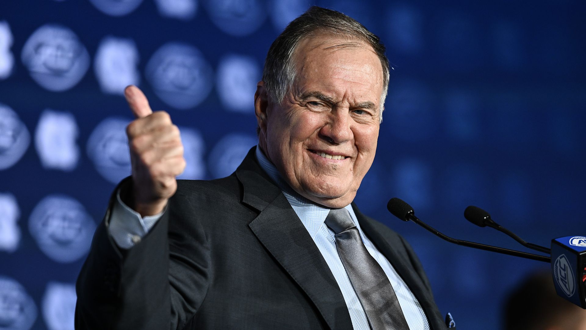 Smiling older man in dark suit and silver tie gives thumbs-up at podium with blue background and microphones displaying ACC logo.