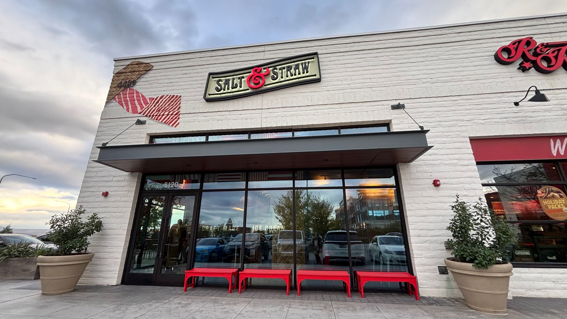 Front view of a white brick Salt & Straw ice cream shop with a lit sign reading "Salt & Straw". A red cone mural decorates the left wall; red benches sit outside under a cloudy sky.
