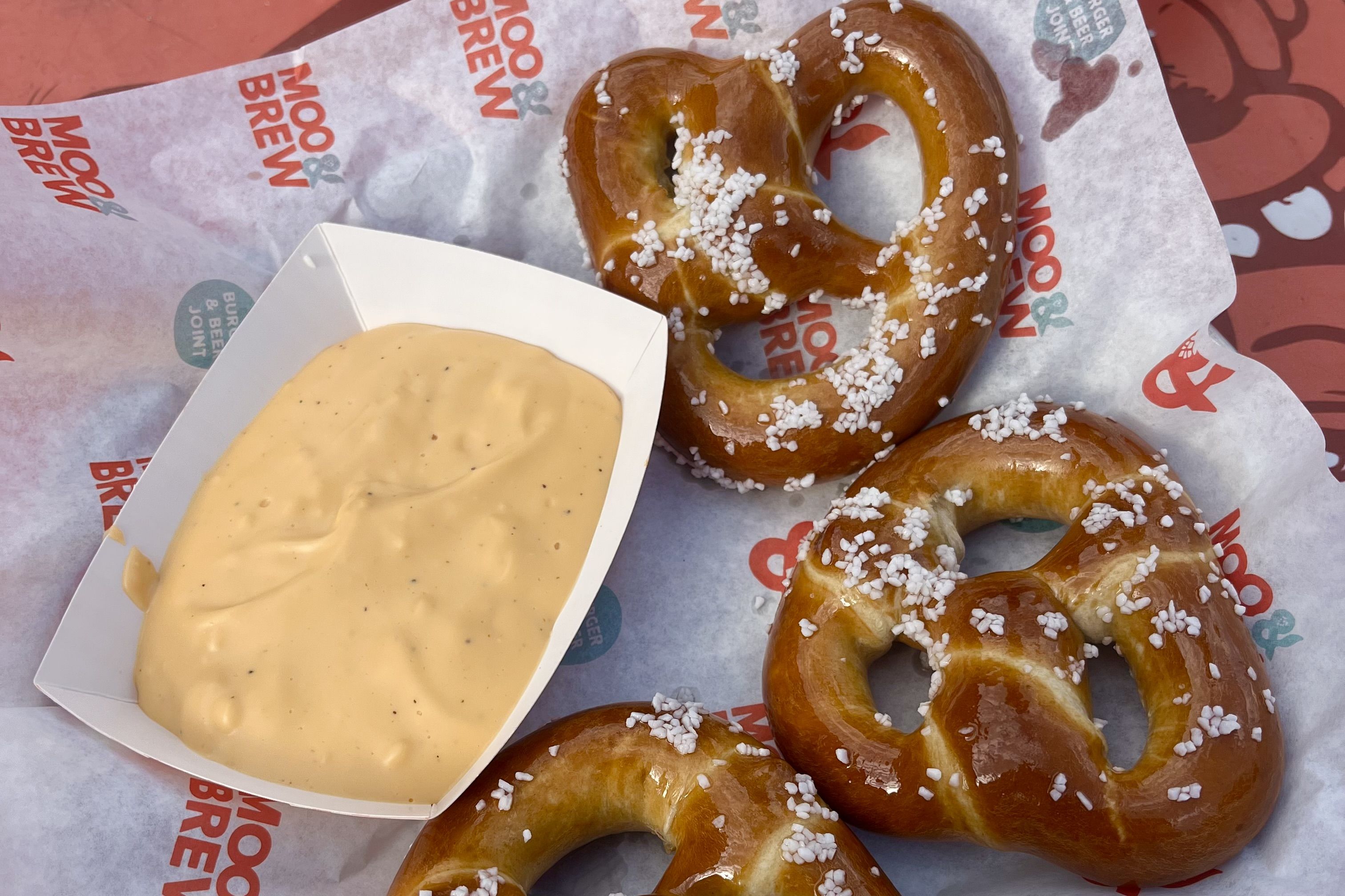 Several glossy pretzels with coarse salt sit on branded parchment next to a pale cheese dipping sauce in a white carton.