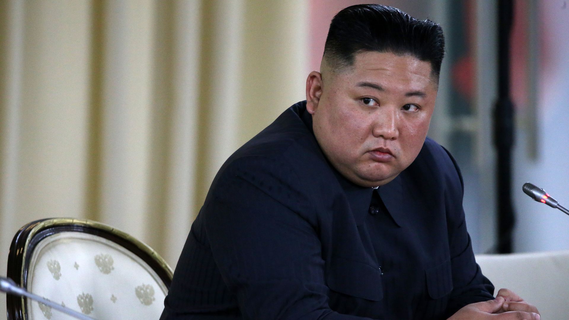 This image shows Kim John Un sitting in front of a microphone and looking to the left.