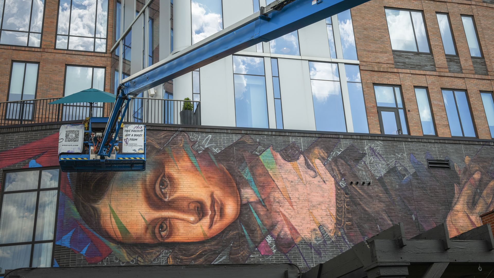 Modern brick-and-glass building with windows reflecting clouds. A blue construction beam spans the frame as a suspended lift sits left, under a colorful mural of Mona Lisa's face on the wall.