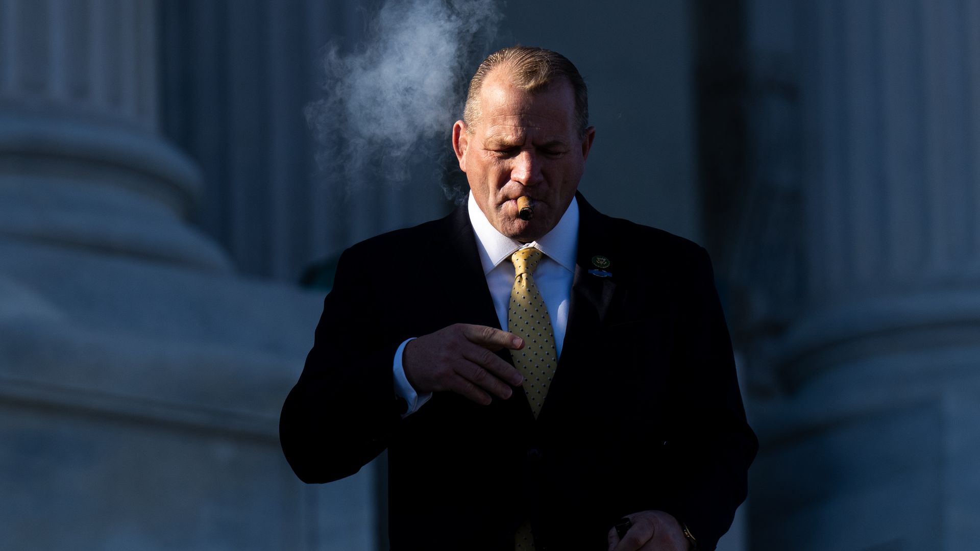 Rep. Troy Nehls, wearing a dark grey suit, white shirt and yellow tie and smoking a cigar with white marble columns in the backdrop.