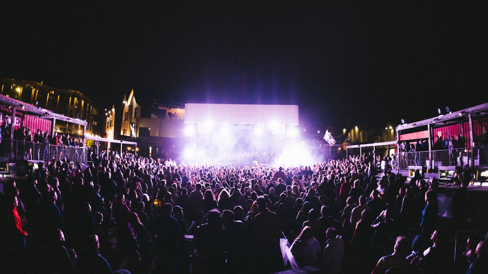 A large crowd on a rooftop at night with bright lights on the stage. 
