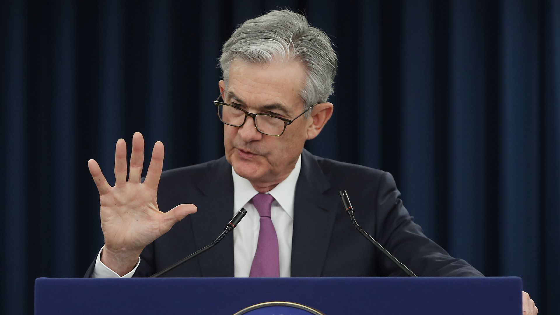 In this image, Powell stands and speaks while gesturing with his palm facing out.