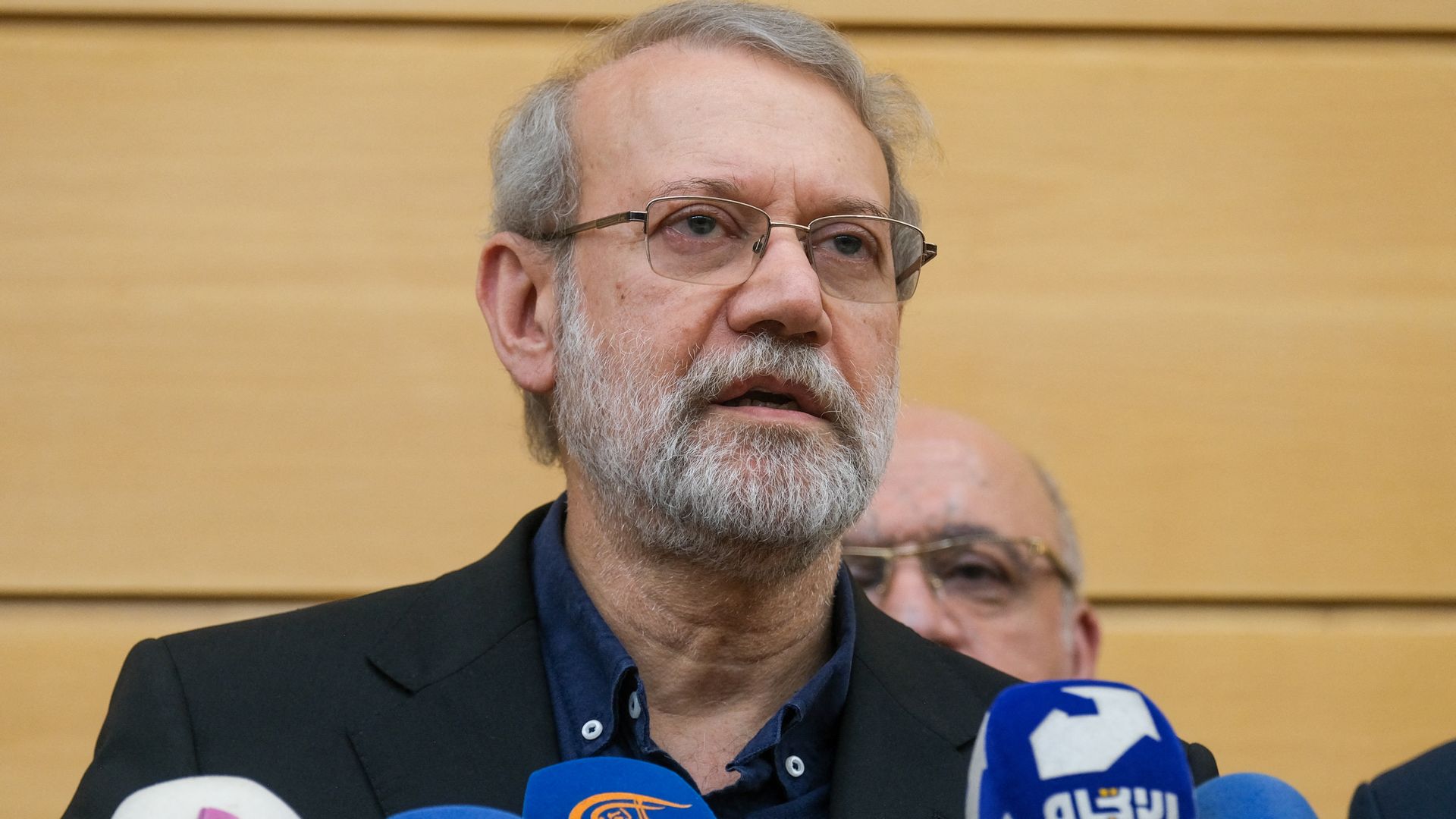 Iranian official Ali Larijani speaks into several microphones during a press briefing in Beirut, with another man standing behind him.