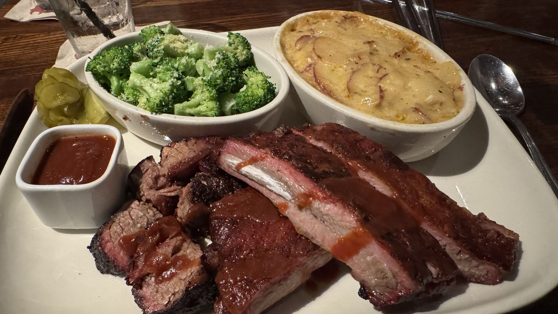 Plate of sliced smoked BBQ ribs with a pink smoke ring, plus broccoli, cheesy au gratin potatoes, and a small dish of red barbecue sauce; pickle slices and a glass of water on a dark wooden table.