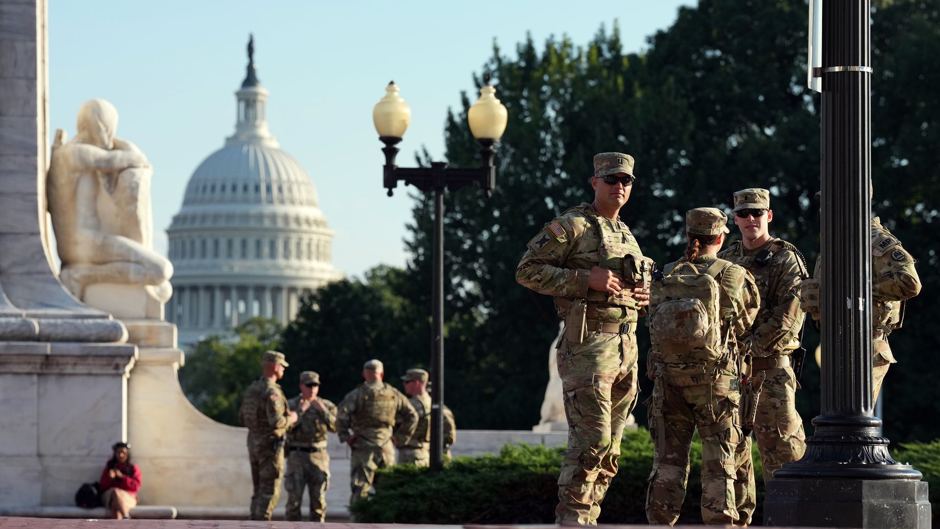 Group of soldiers in camouflage uniforms standing and talking near a marble statue, with the U.S. Capitol dome visible in the background under clear sky.