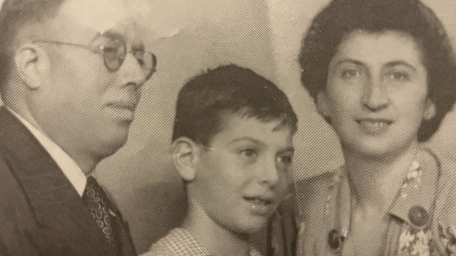 Sepia-toned vintage group portrait showing a man with glasses on the left, a boy in front, and a smiling woman with dark, curly hair on the right, all in formal attire.