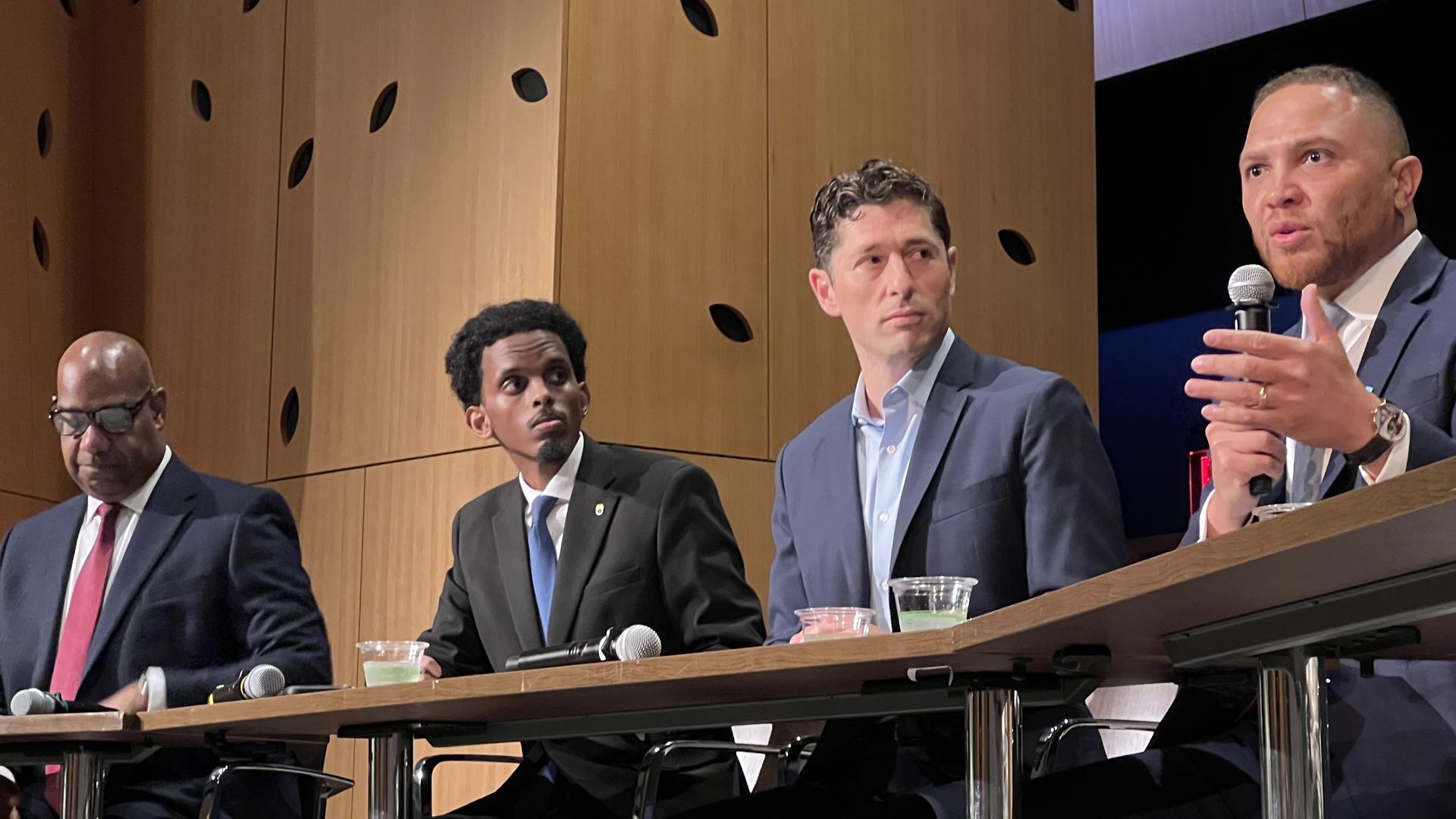 Four men in suits sit at a panel table in a modern room with wooden walls. One man on the right speaks into a microphone while gesturing with his hand.