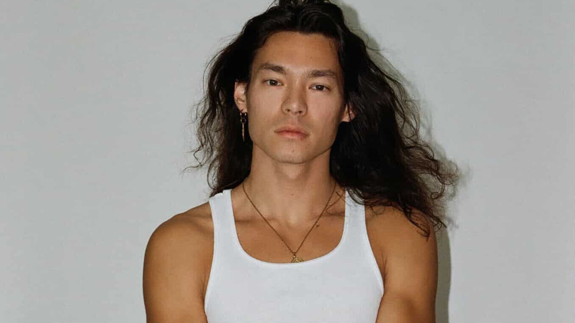 Young man with long dark hair wearing a white tank top and light blue jeans stands against a plain gray background with one arm crossed over the other.
