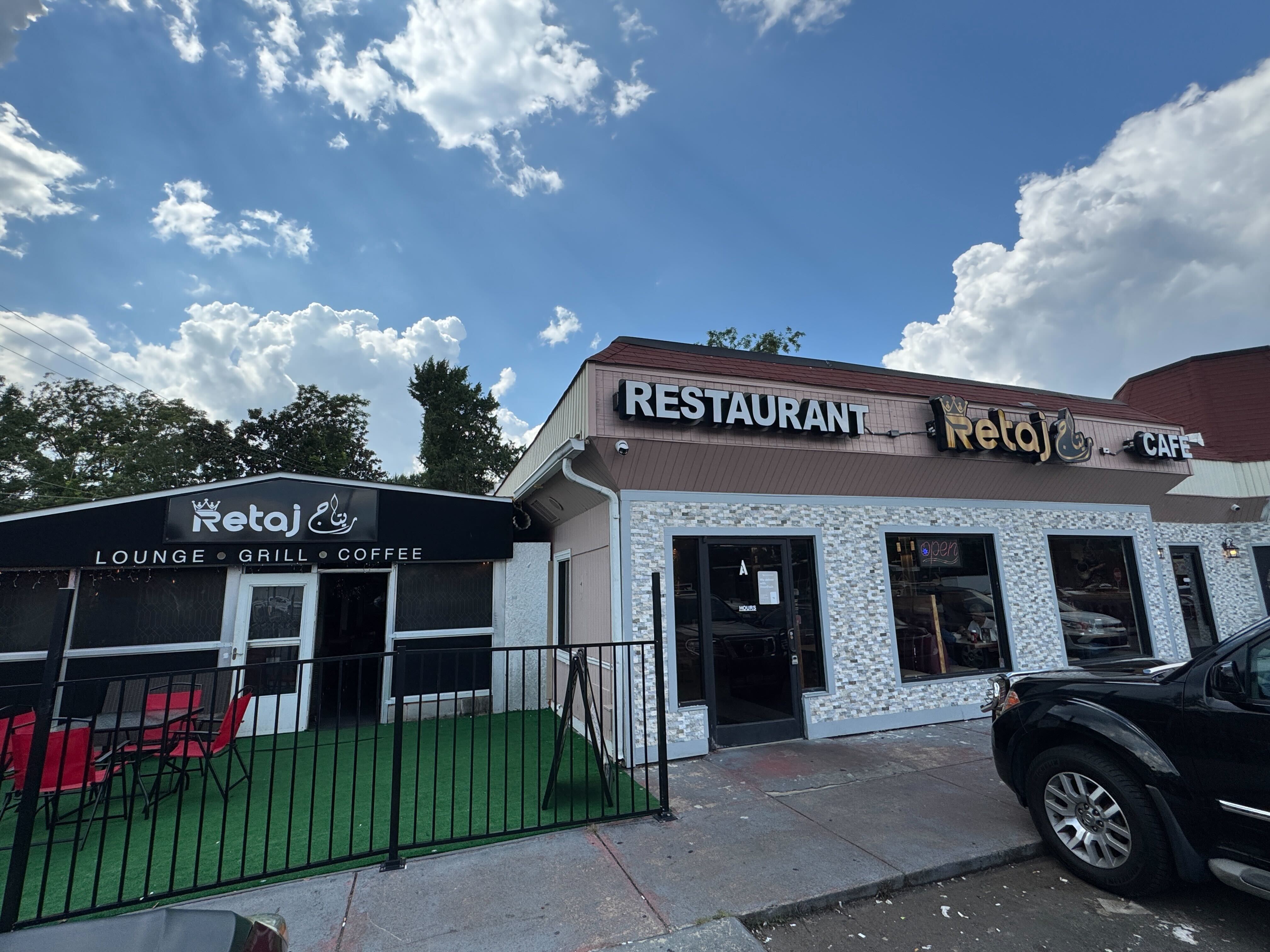 Exterior of Retaj restaurant and cafe with black and white signs, red patio chairs, and a green artificial grass seating area under a blue sky with clouds.