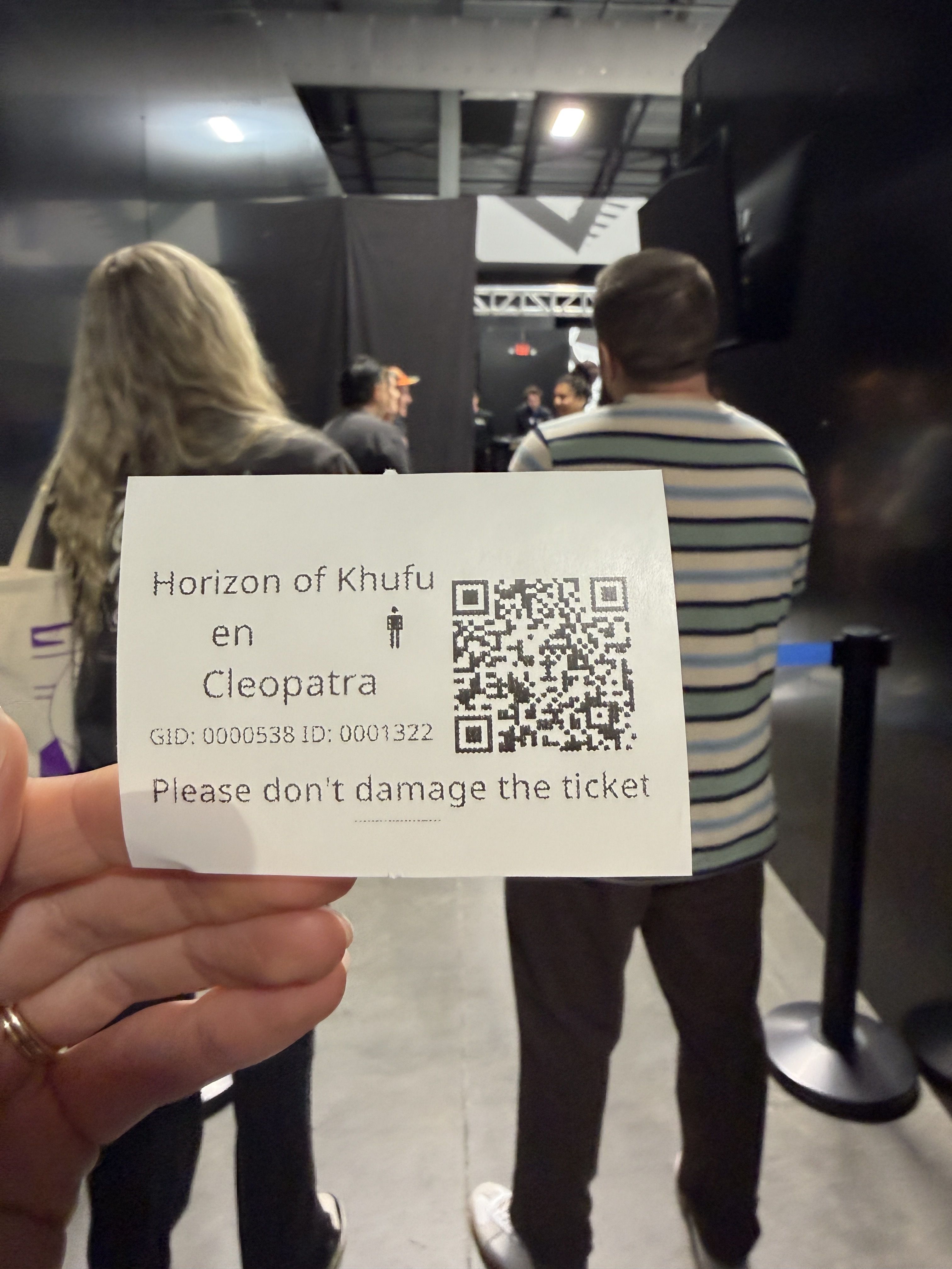 Close-up of a hand holding a white museum ticket with a QR code. It reads "Horizon of Khufu en Cleopatra" and "Please don't damage the ticket." In the background, people queue near black exhibit walls.