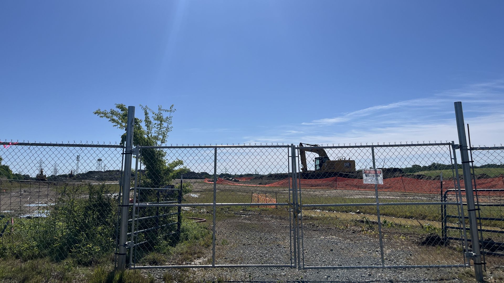 Blue sky with sun glare over a chain-link fence at a construction site. Orange safety netting along the ground; a yellow excavator behind the fence, with sparse vegetation and a few trees.