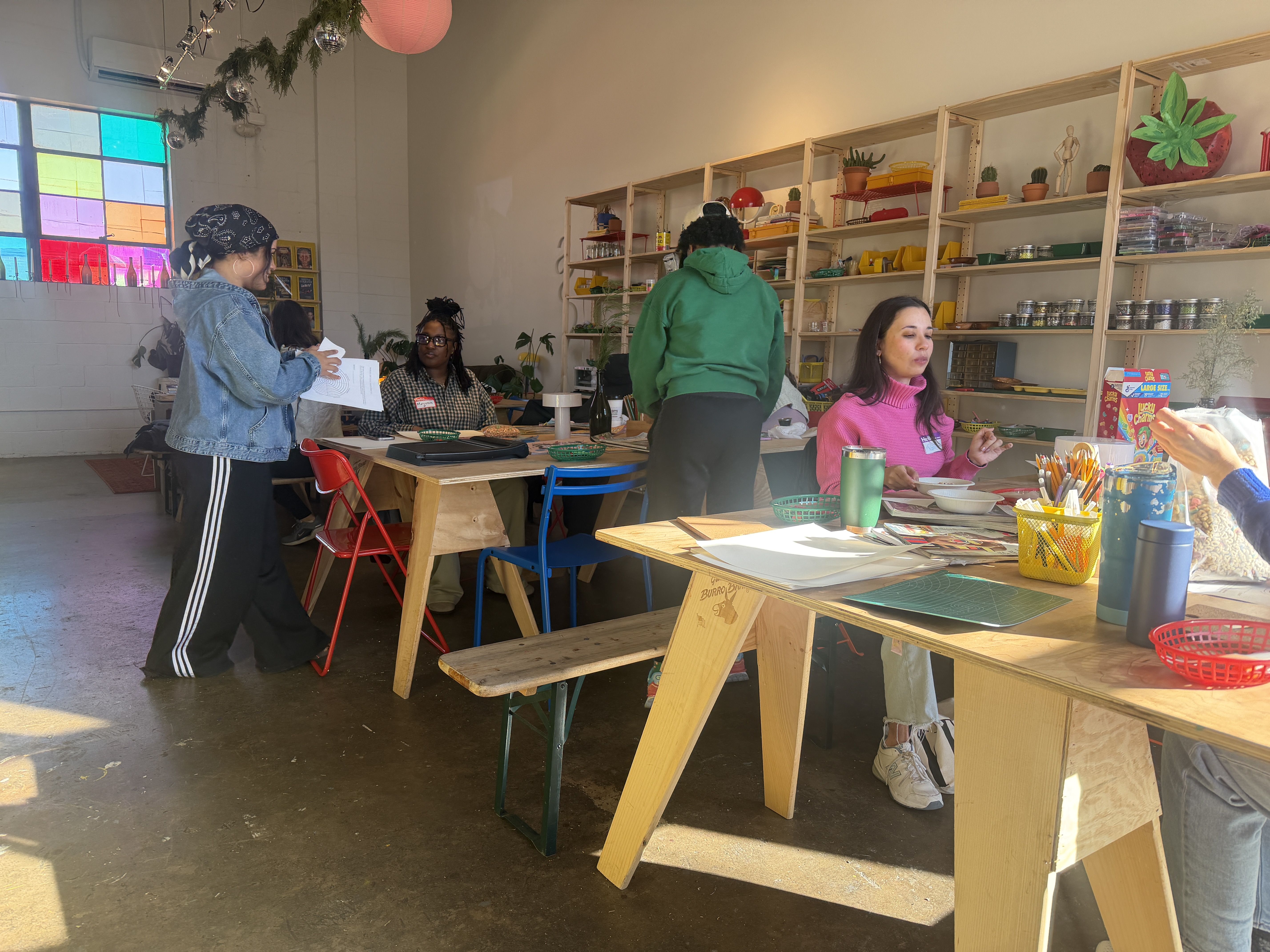 People engaged in creative work in a brightly lit room with wooden tables and shelves filled with craft supplies. A large colorful stained-glass window casts light inside.