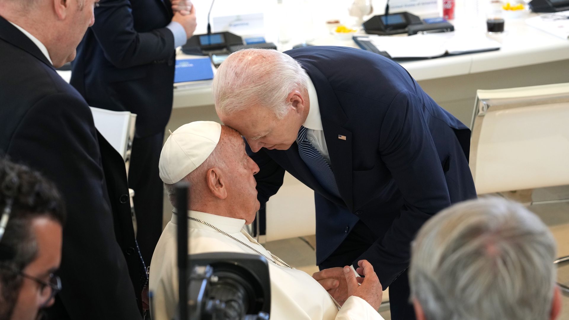 Pope Francis is seated as President Biden lowers his forehead to the pope's. 