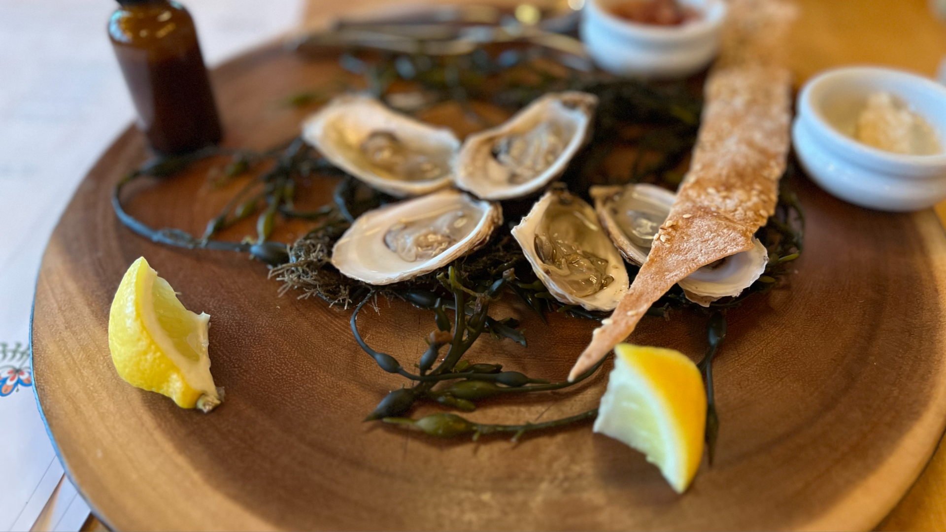 Oysters on their half-shells served on a wooden platter with a piece of flat crust and lemons on a bed of seaweed.