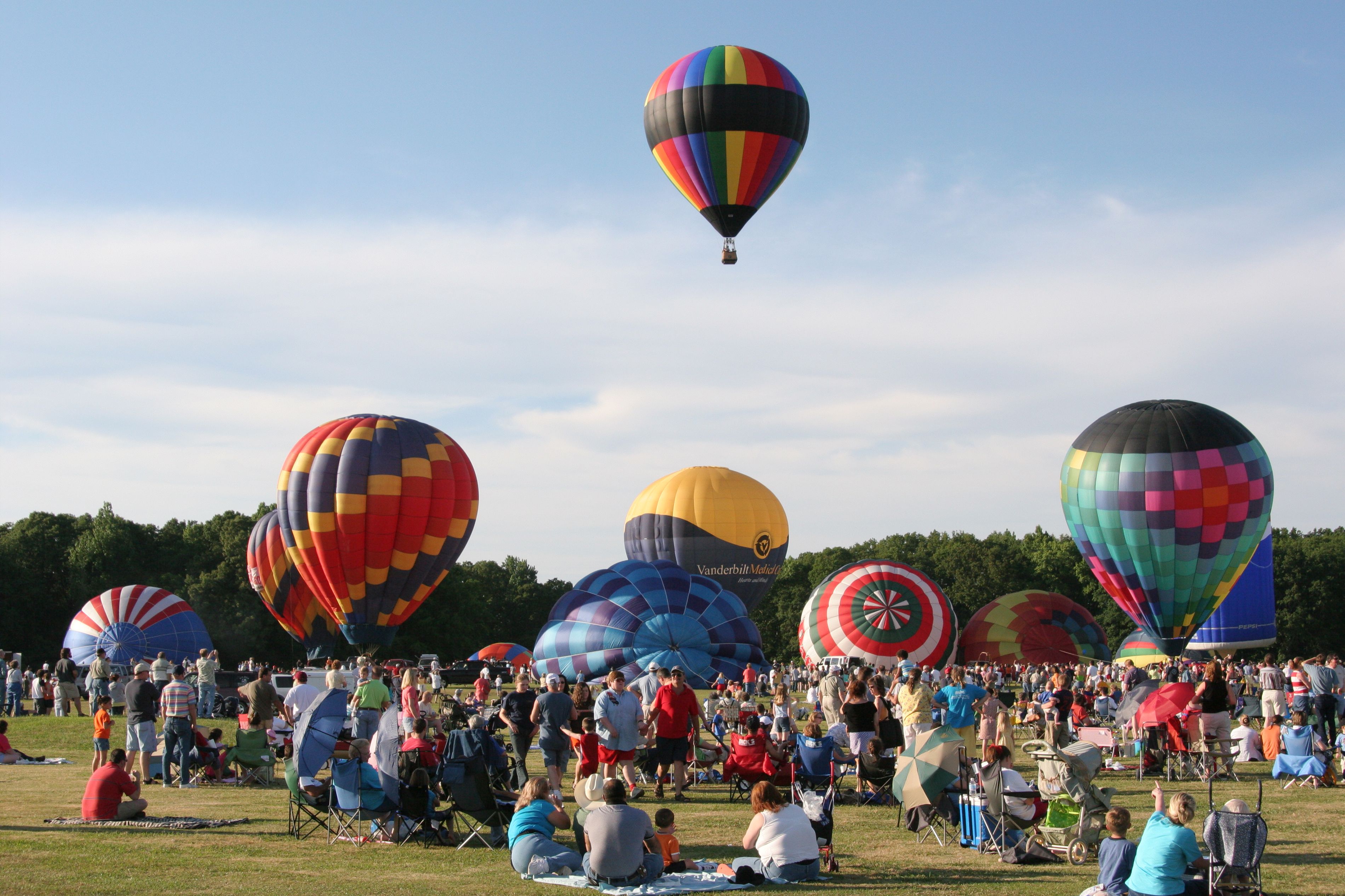 A crowd is shown in a field as multiple, colorful hot air balloons fill up and float away.