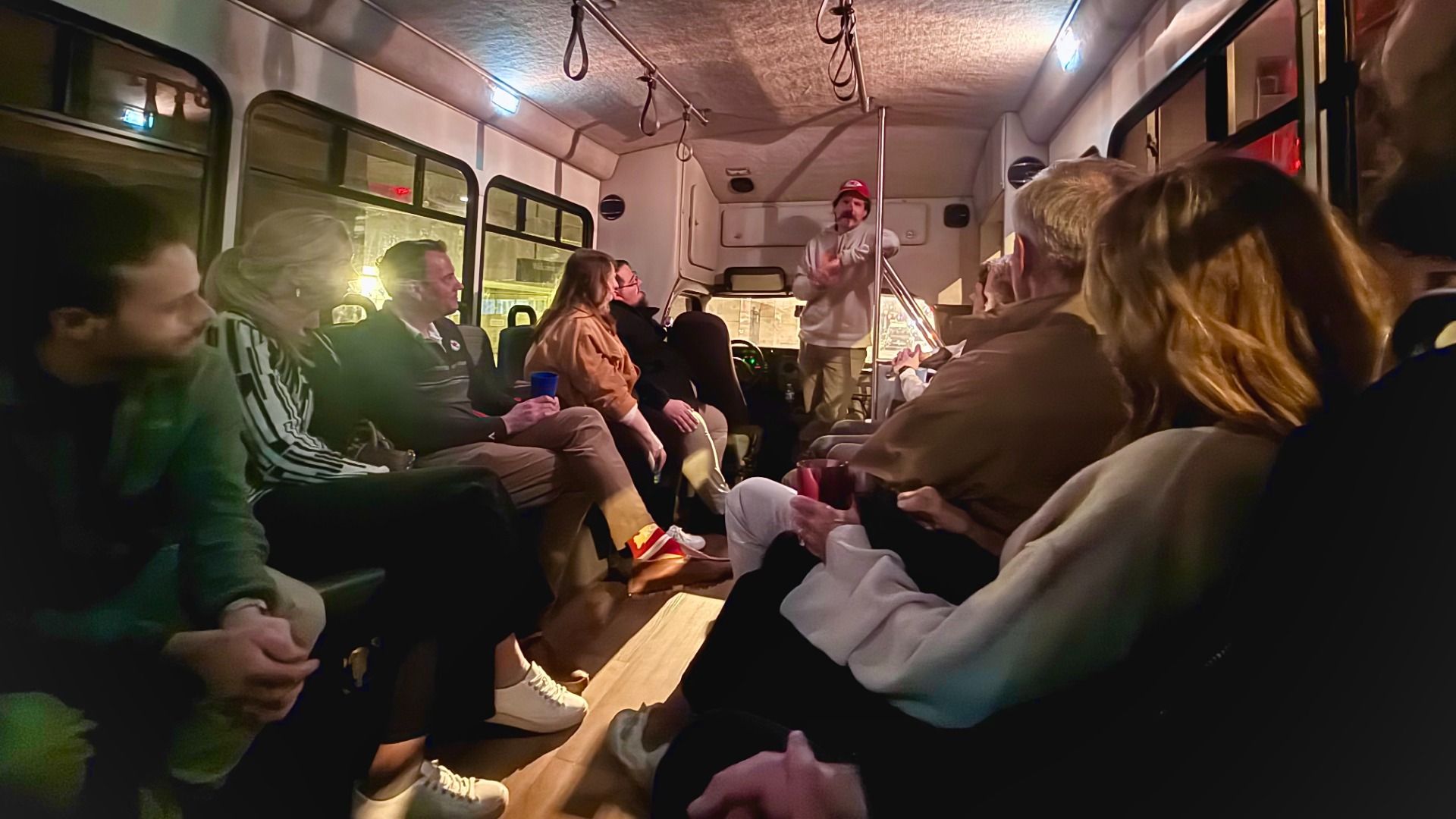 Group of people seated inside a dimly lit bus listening to a man in a white sweatshirt and red cap standing at the front, gesturing with his hands during a nighttime event.