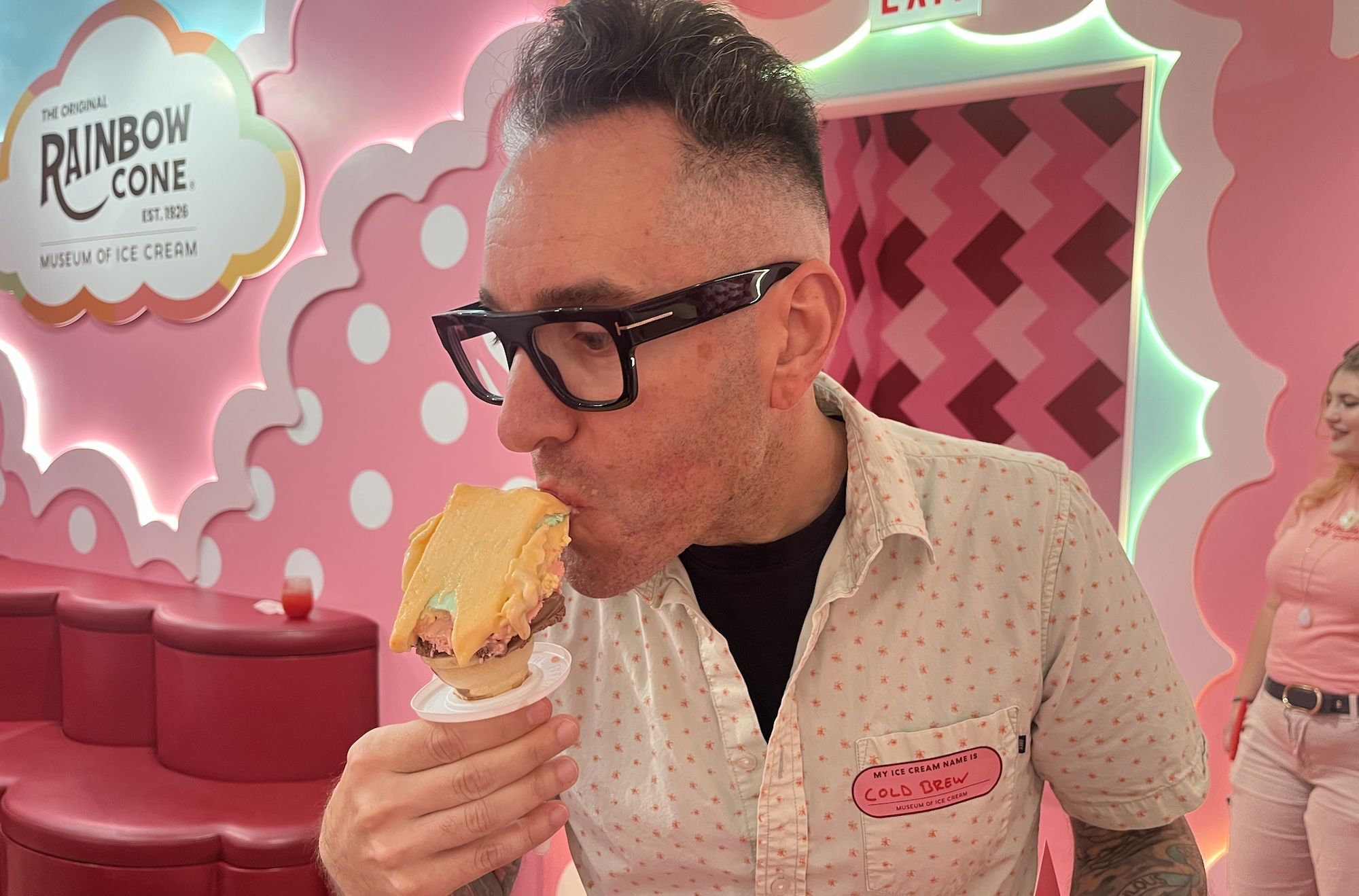 Man with thick black glasses and a short mohawk eats a giant multi-scoop ice cream cone in a pink, retro Rainbow Cone shop, with pastel walls and bold signage in the background.