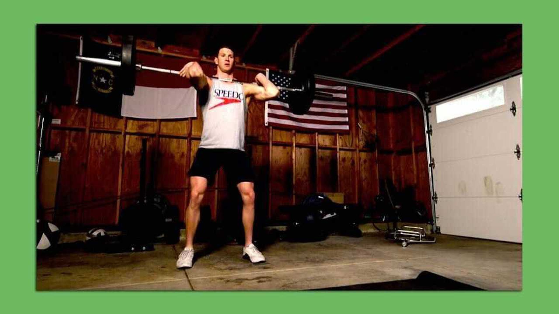 A man in a white t-shirt presses a barbell in a dark room in front of a wooden wall with flags