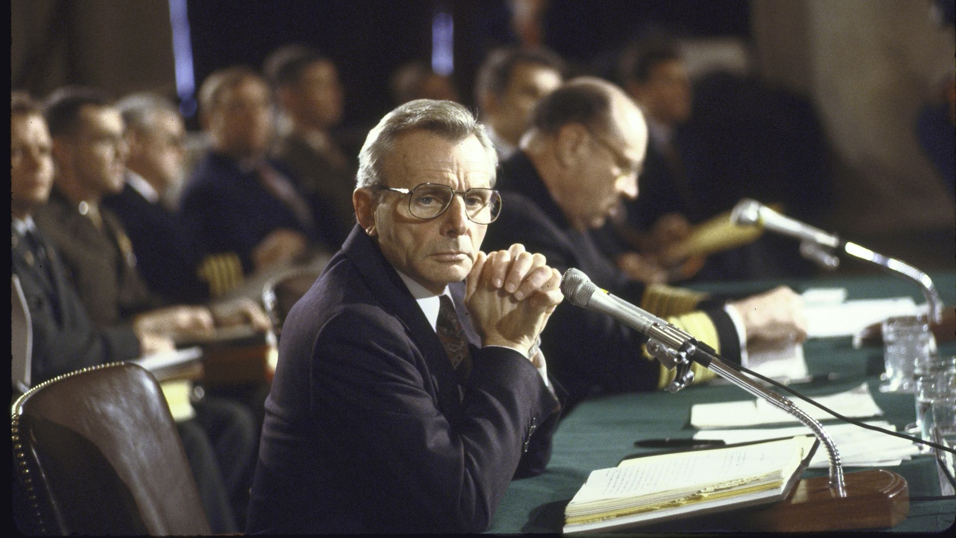 Frank Carlucci sits with hands steepled at a wooden table, looking towards the camera sideways.