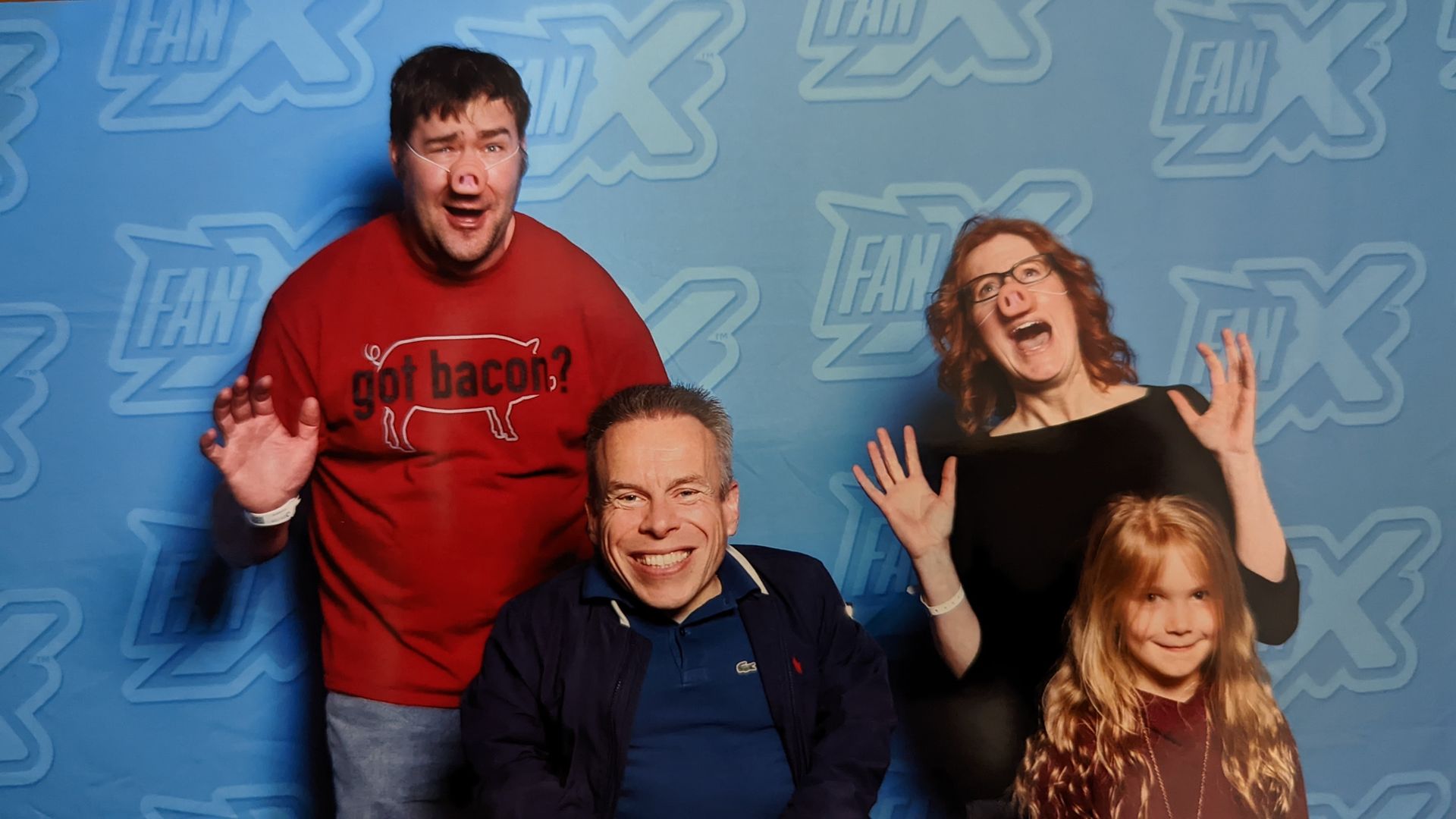 A man and woman in fake pig noses pose with their daughter for a photo with actor Warwick Davis.
