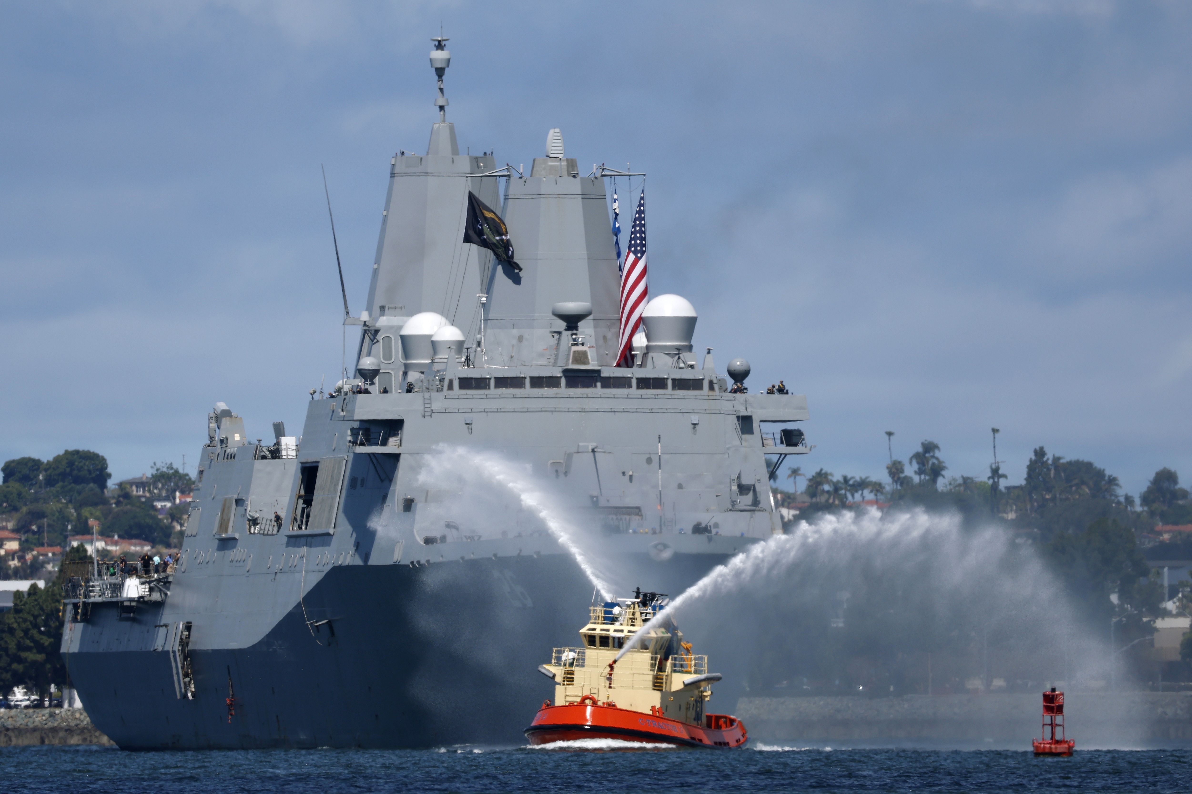 A large U.S. Navy warship sails forward as a small tugboat in front sprays arcing streams of water, with an American flag flying from the ship and a shoreline visible in the background.