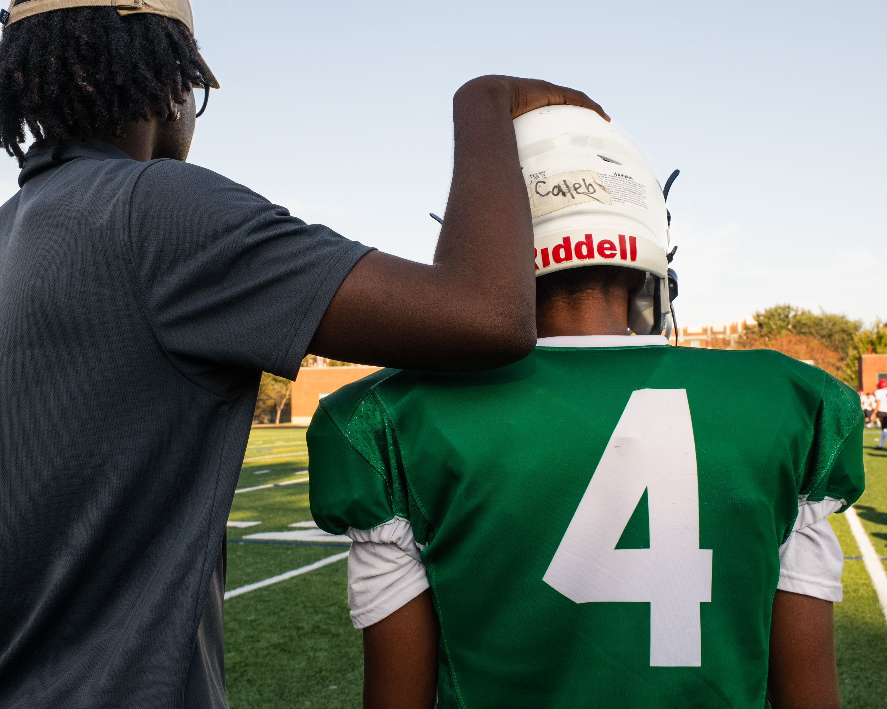 A coach in gray shirt and beige cap places a hand on the helmet of a football player wearing a green jersey with white number 4, named Caleb, standing on a sunny field.