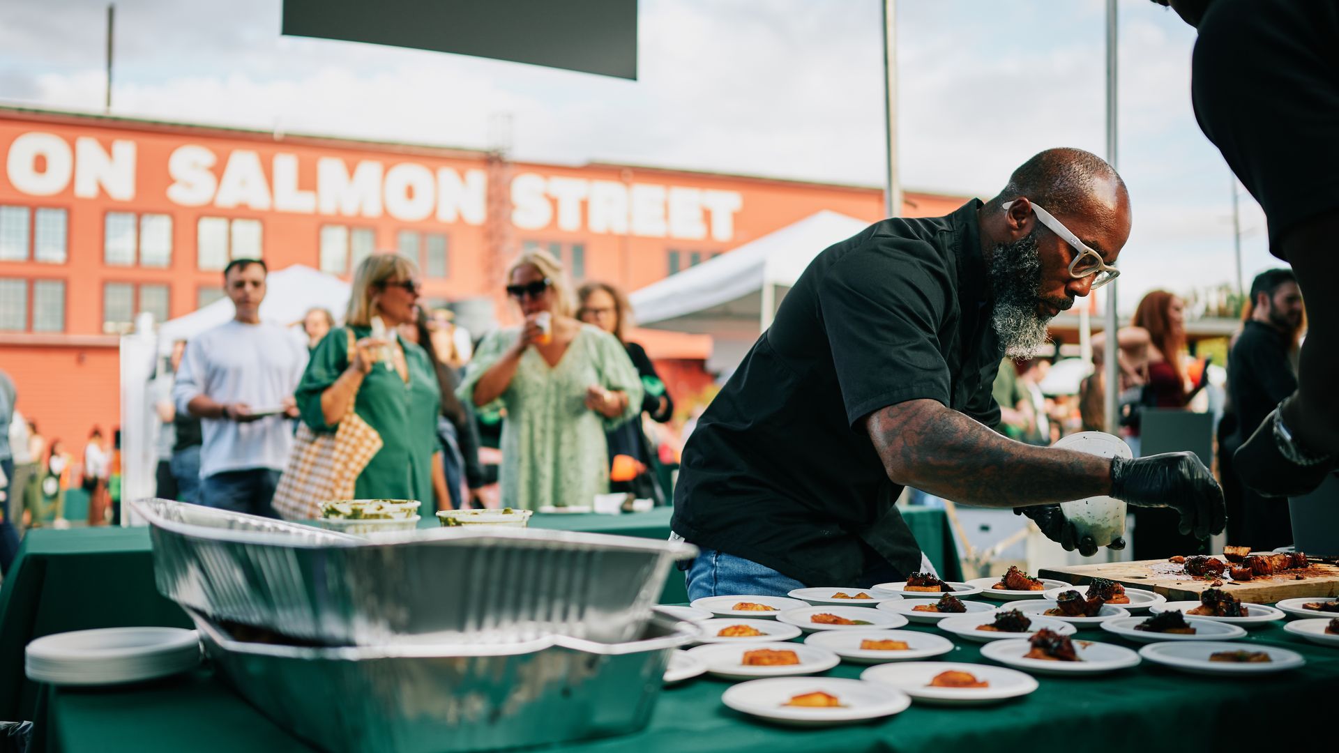Bearded man in a black shirt grills meat at an outdoor food event, while a crowd watches; plates on a green table, and a red brick building reads ON SALMON STREET in the background.