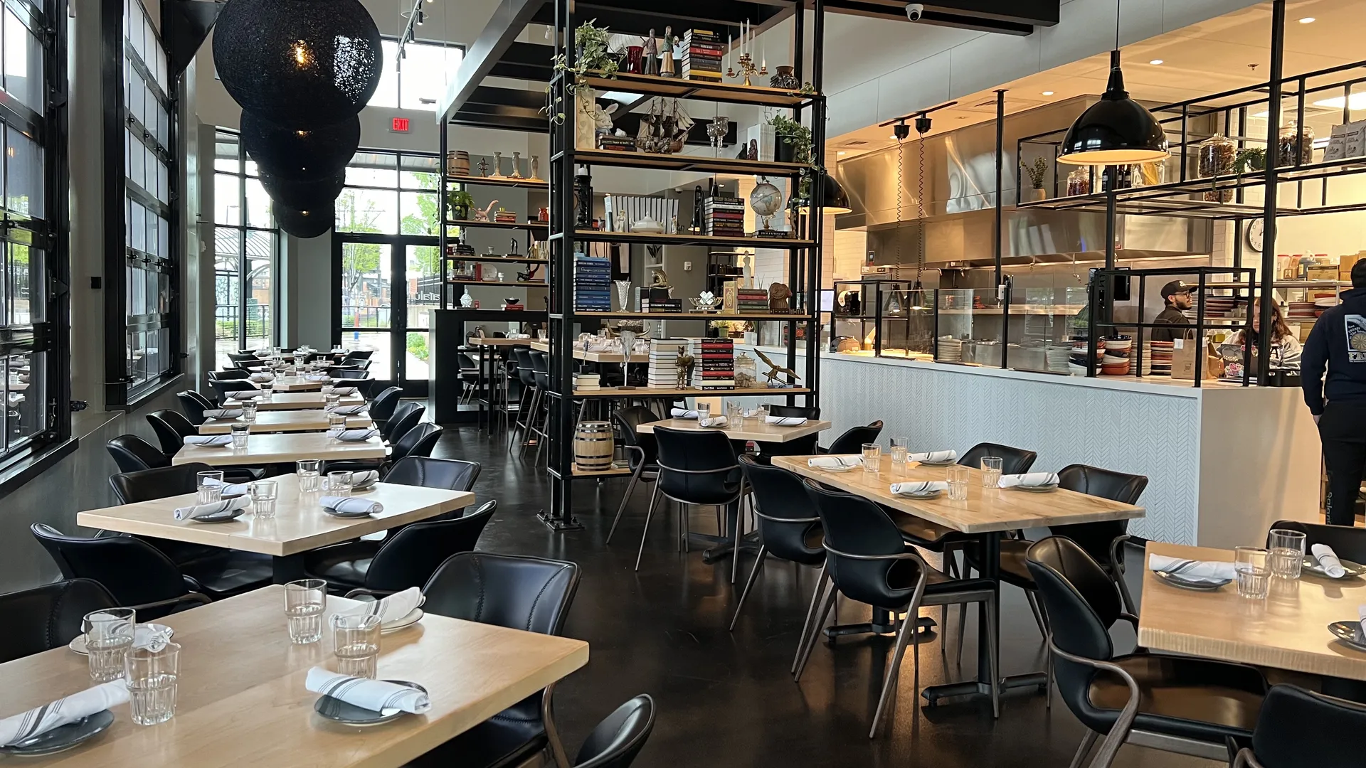 Modern restaurant interior with natural wood tables, black chairs, glassware, and rolled white napkins. Large windows, open kitchen, and decorative shelves with books and plants.