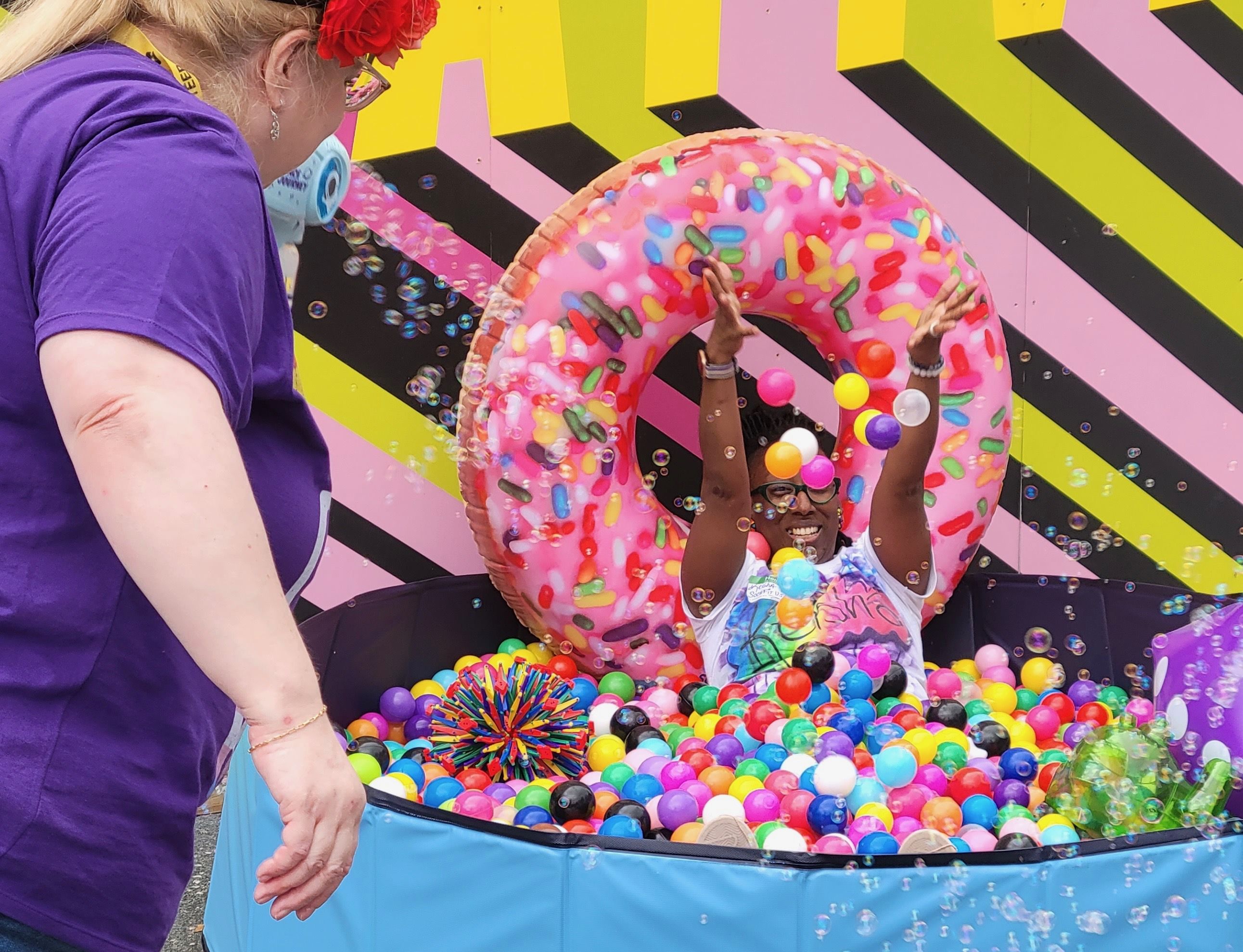 Person in colorful ball pit with inflatable pink donut, throwing colorful balls up joyfully. Another person in purple shirt with red flower in hair is nearby. Background is pink, yellow, black stripes.