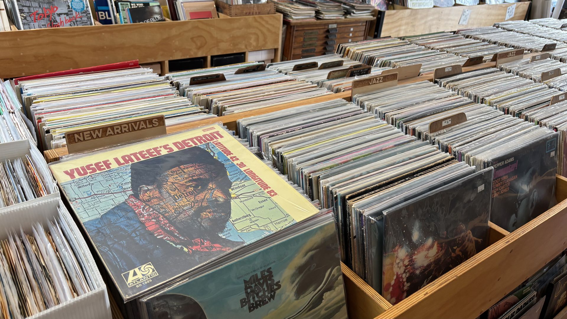 Rows of vinyl records in wooden bins at a record store, featuring a prominent album titled "Yusef Lateef's Detroit" under a "New Arrivals" sign, with sections labeled Jazz and others.