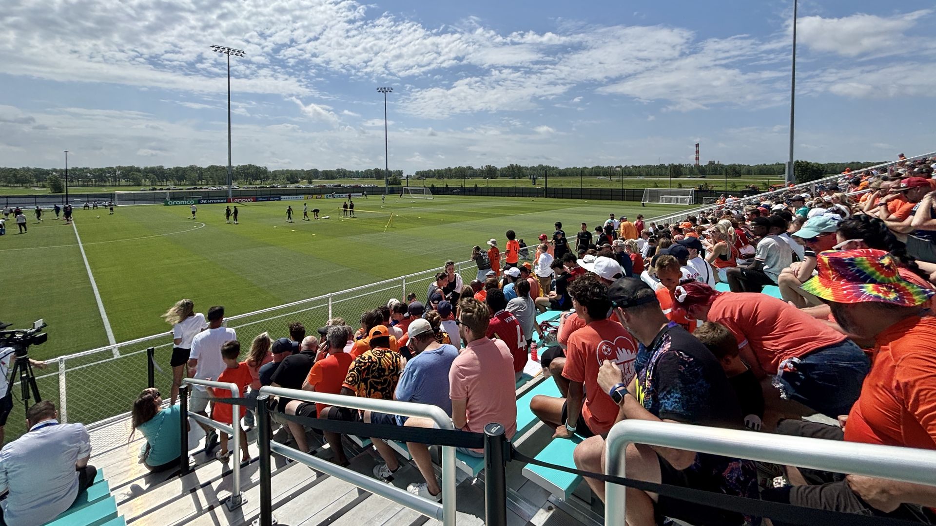Sunny day at a soccer stadium. Crowds in red seat on the right as players run drills on the green field near goalposts, with tall floodlights and a bright blue sky.