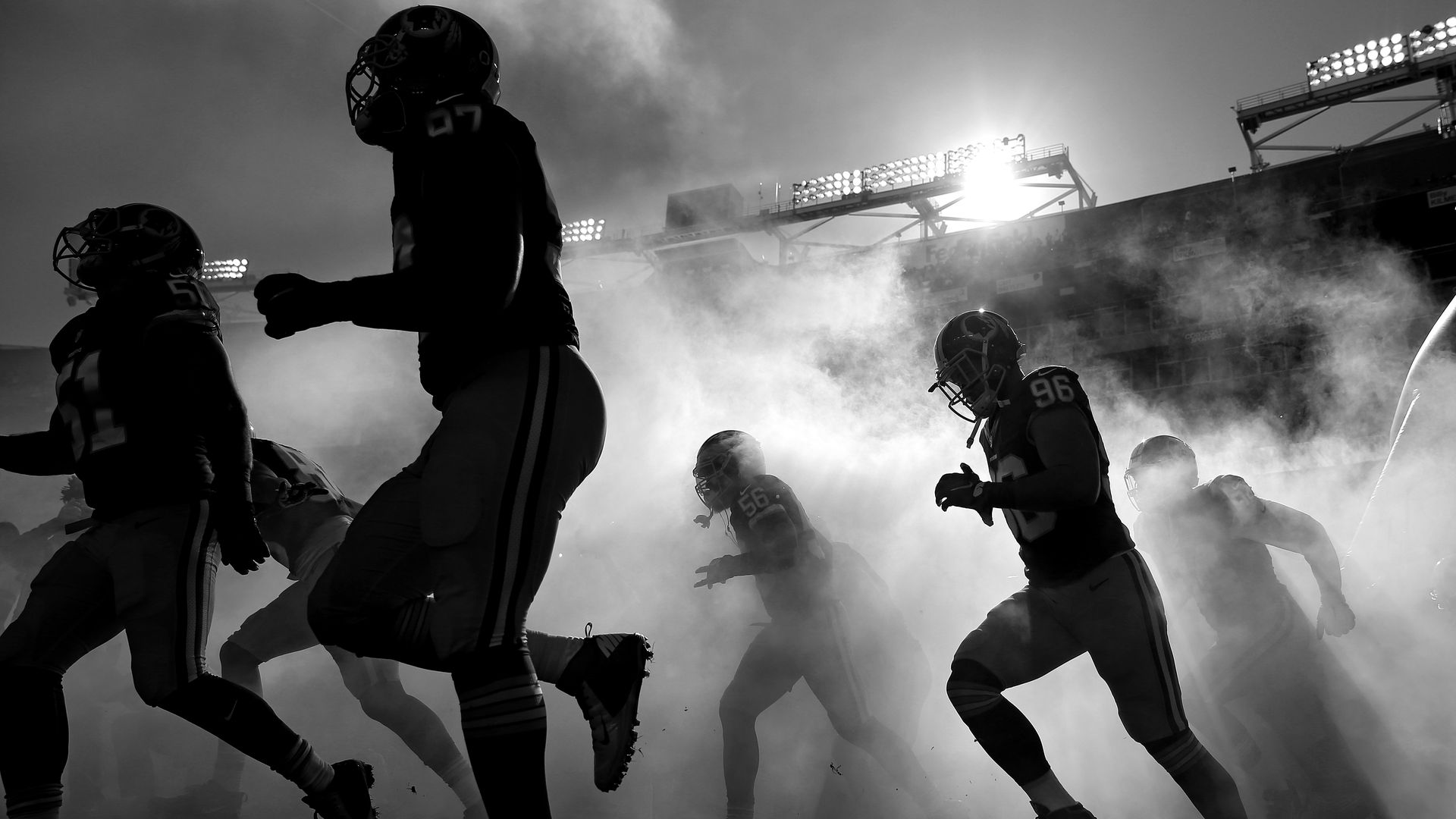 A black and white image: Washington football players run onto the field through a haze of fog.