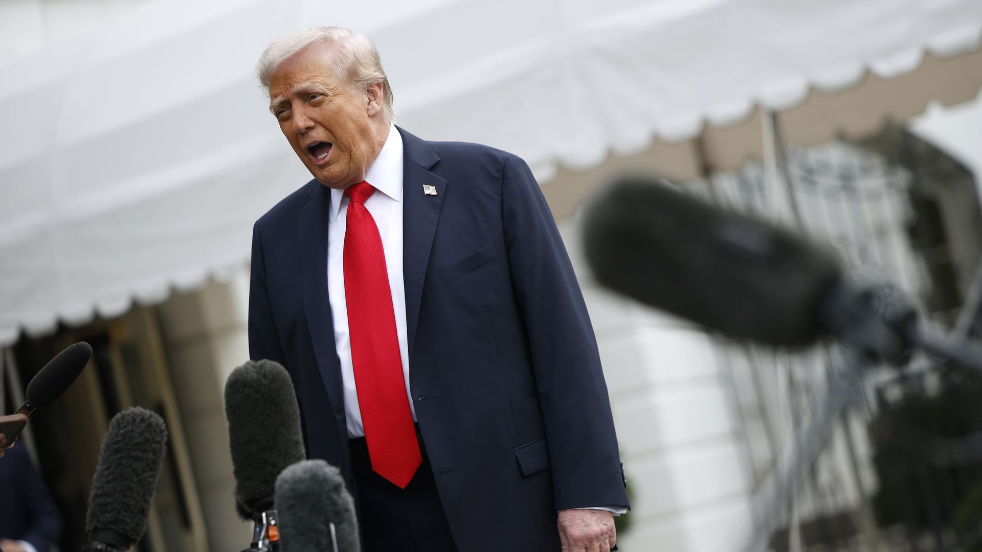 Photo of a man standing taking questions from reporters while he is in a suit on a lawn.