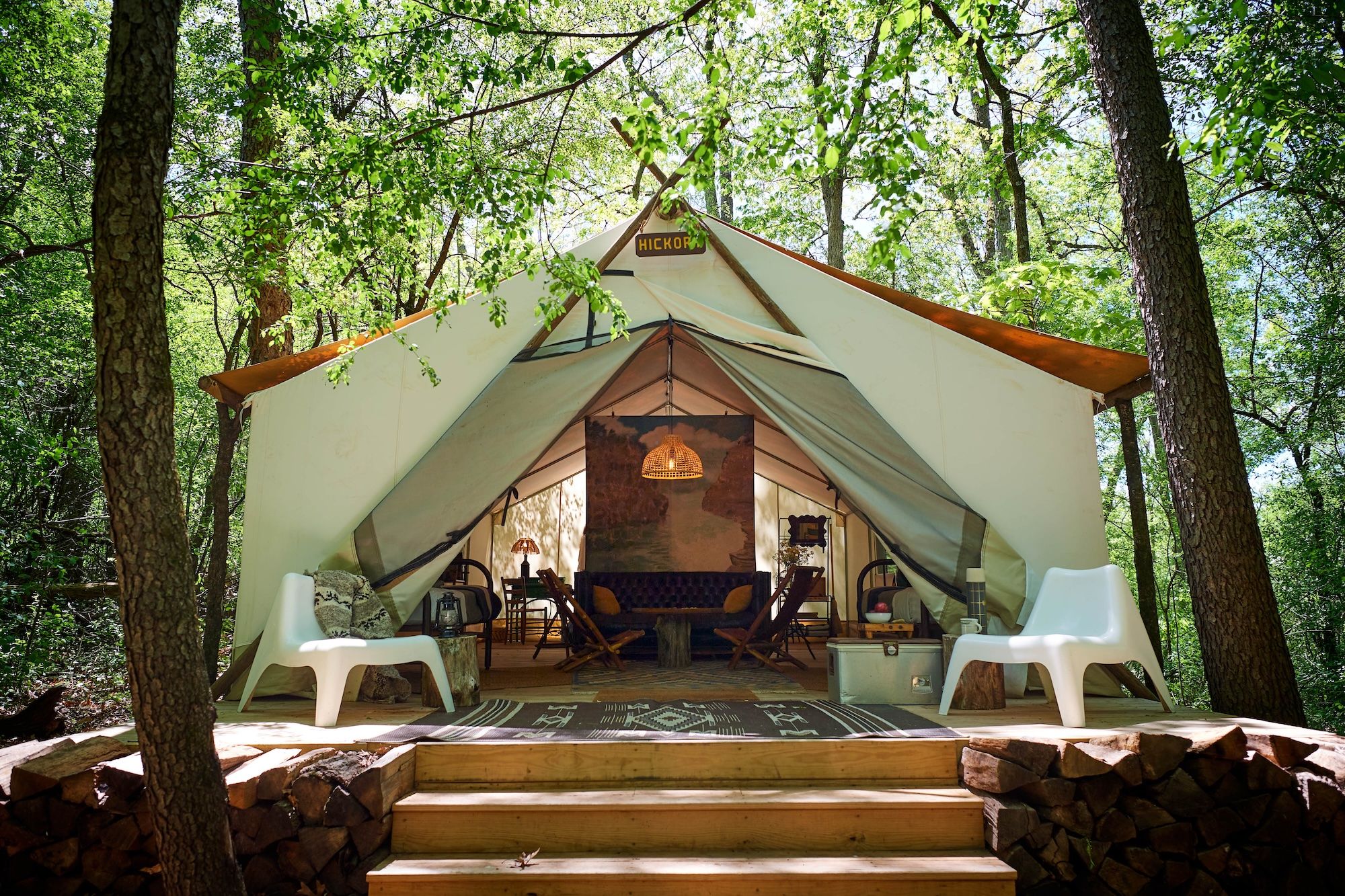 A white tent in the woods with two white chairs on the touside and chairs and a couch inside.